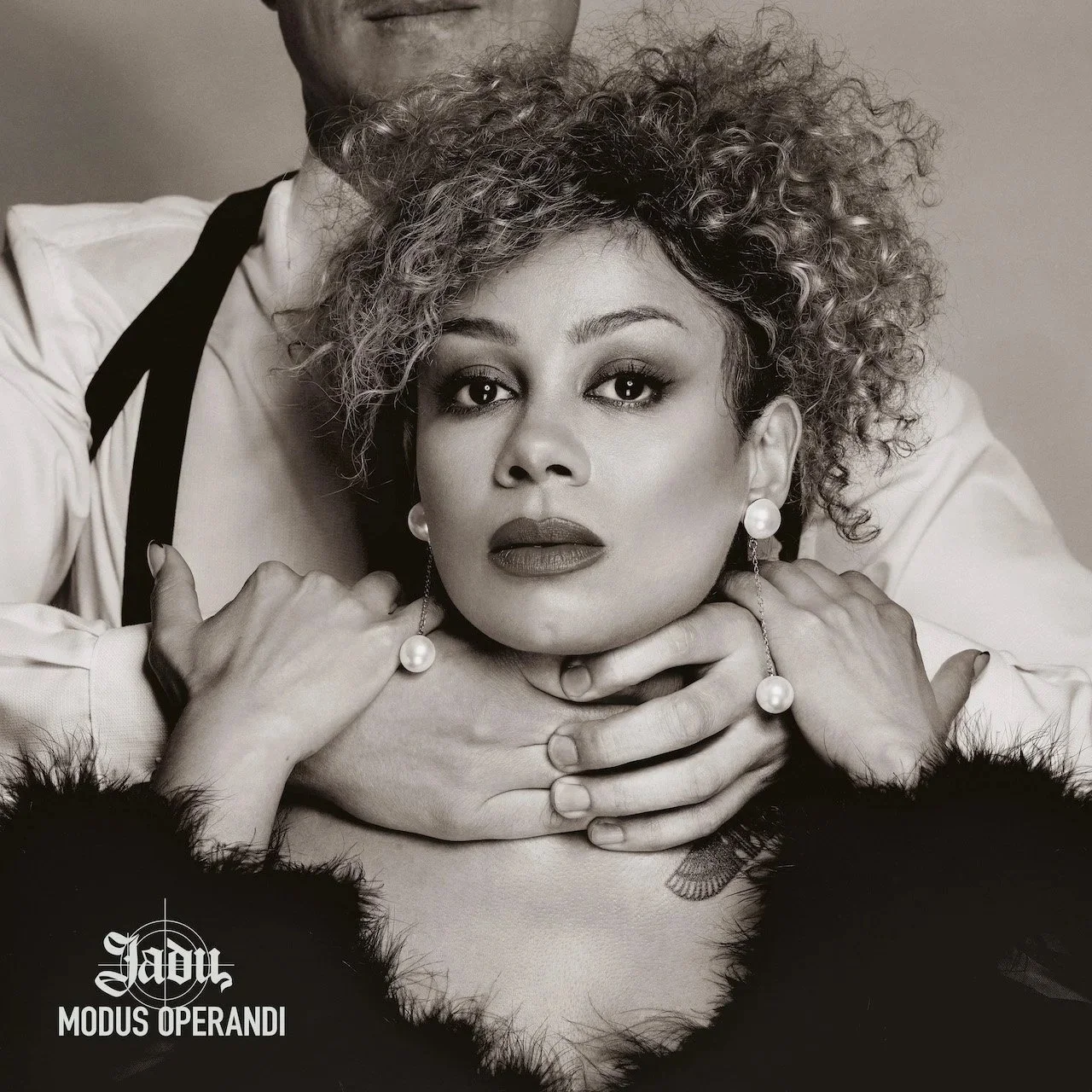 Schwarzweiß-Fotografie der Künstlerin mit lockigen Haaren und großen Perlenohrringen, das von einem Mann im Hintergrund mit einem weißen Hemd und schwarzen Hosenträgern umarmt wird.