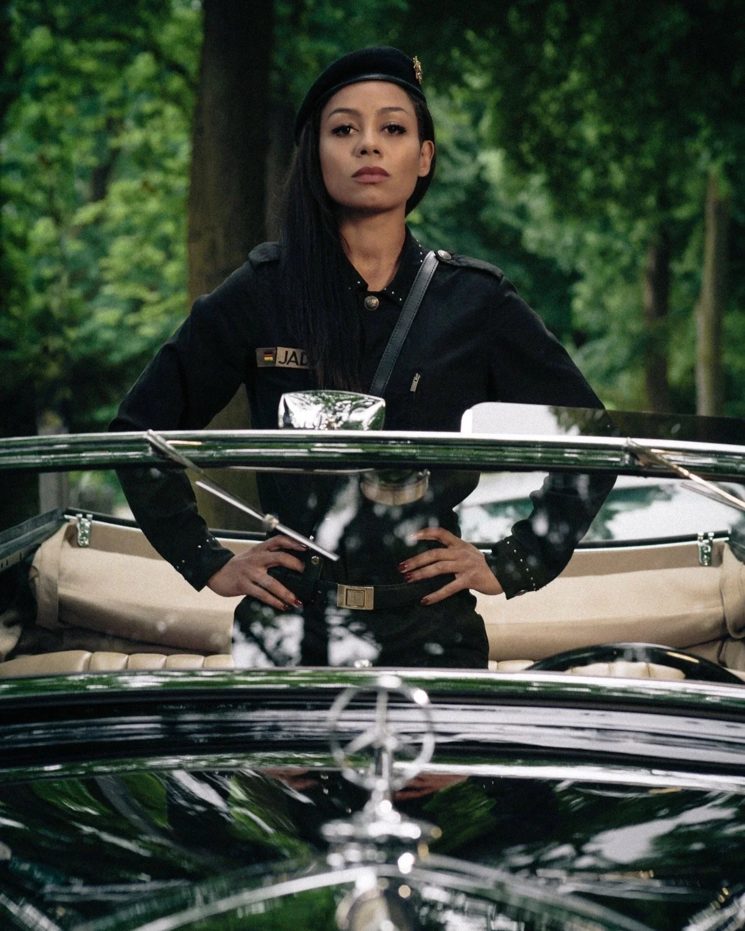 Eine Frau in einer schwarzen Uniform steht hinter einem klassischen Mercedes-Benz Auto in einem grünen Wald. Sie hat dunkle Haare, trägt eine schwarze Mütze und blickt ernst.