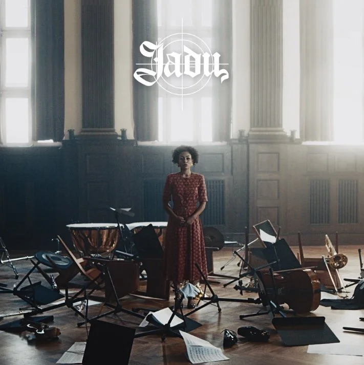 Eine Frau steht in einem Saal mit einem zerstörten Orchester, zurückhaltend in einem Kleid. Über ihr schwebt das Wort 'Jadu' in weißer Schrift.