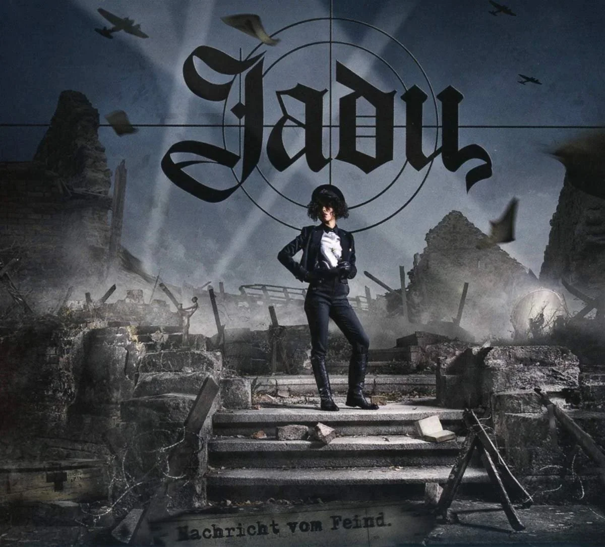 Eine Frau steht auf einer zerstörten Treppe vor einer Wand mit der Aufschrift 'Jadu'. Die Szenerie erinnert an Kriegsschauplätze mit Trümmern und zerrissener Infrastruktur, während kleine Flugzeuge am Himmel fliegen.