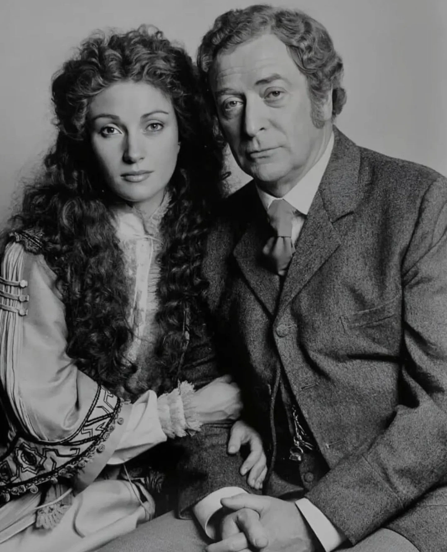 Black and white photo of a young woman with long, curly hair and an older man in a suit, sitting close together and looking at the camera.