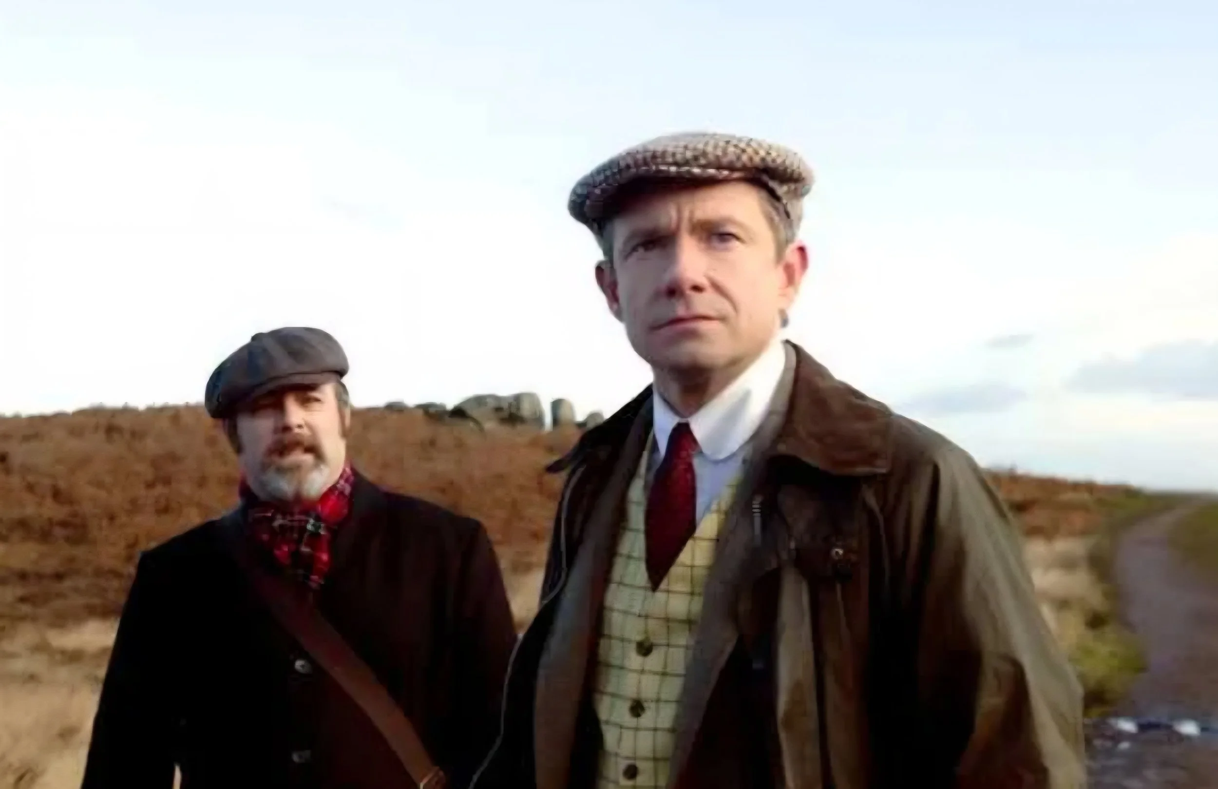 Two men dressed in vintage outdoor clothing standing outdoors on a dirt path with a landscape of grass and rocks in the background.