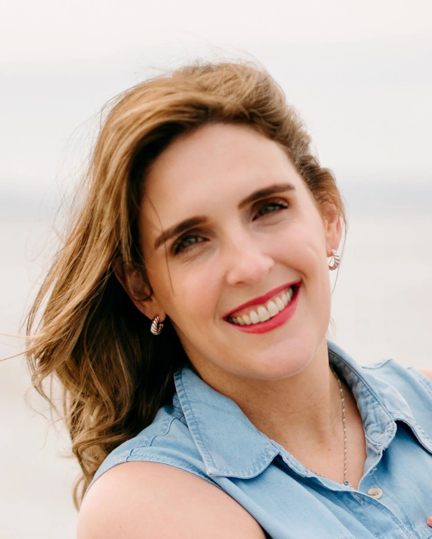 Close-up portrait of a smiling woman with wavy light brown hair wearing a sleeveless denim shirt, outdoors with cloudy sky background.