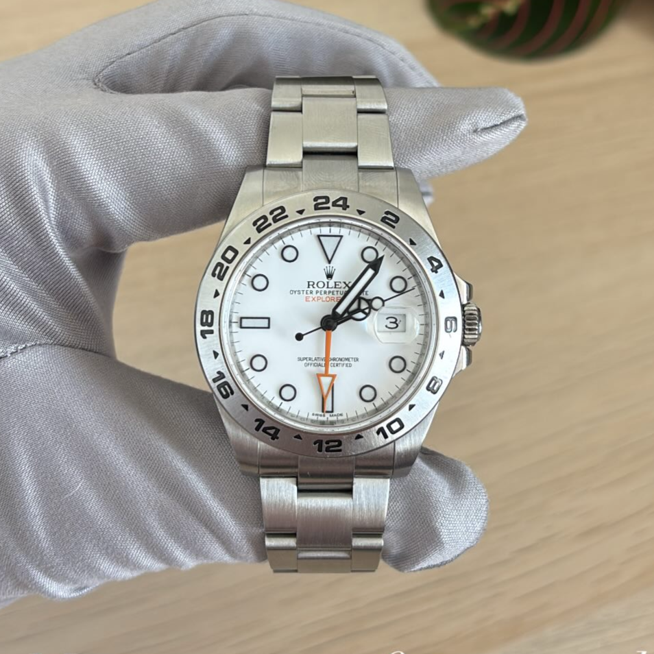 A silver Rolex Oyster Perpetual Explorer II wristwatch with a white dial, black 24-hour bezel, Arabic numerals, date window at 3 o'clock, and a stainless steel bracelet, held by a person wearing a white glove.