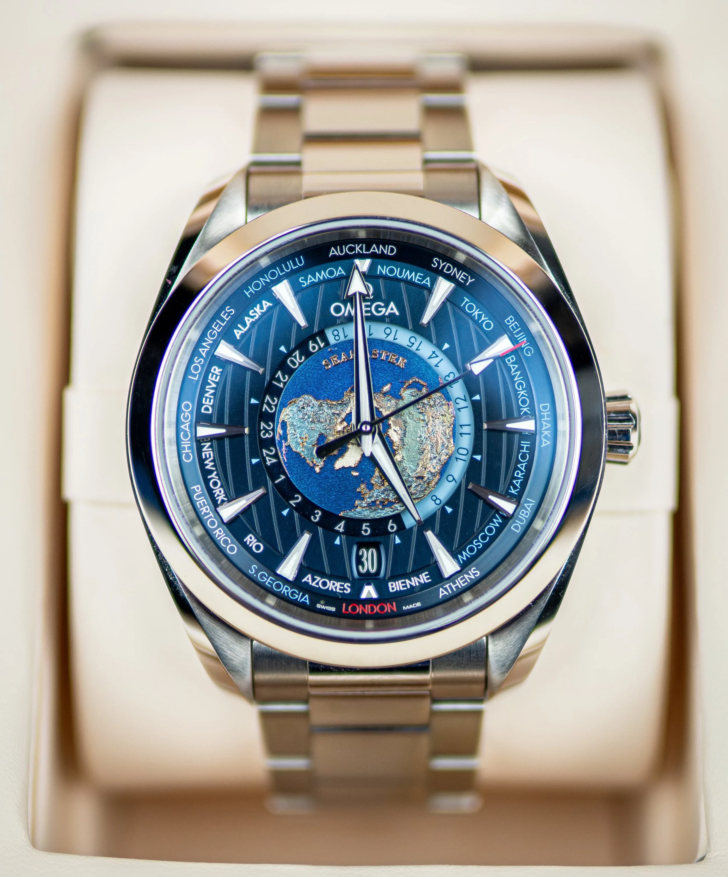 A close-up view of an Omega Seamaster wristwatch with a world map on the dial, displaying time zones for various cities, with a metallic band and a beige watch box.