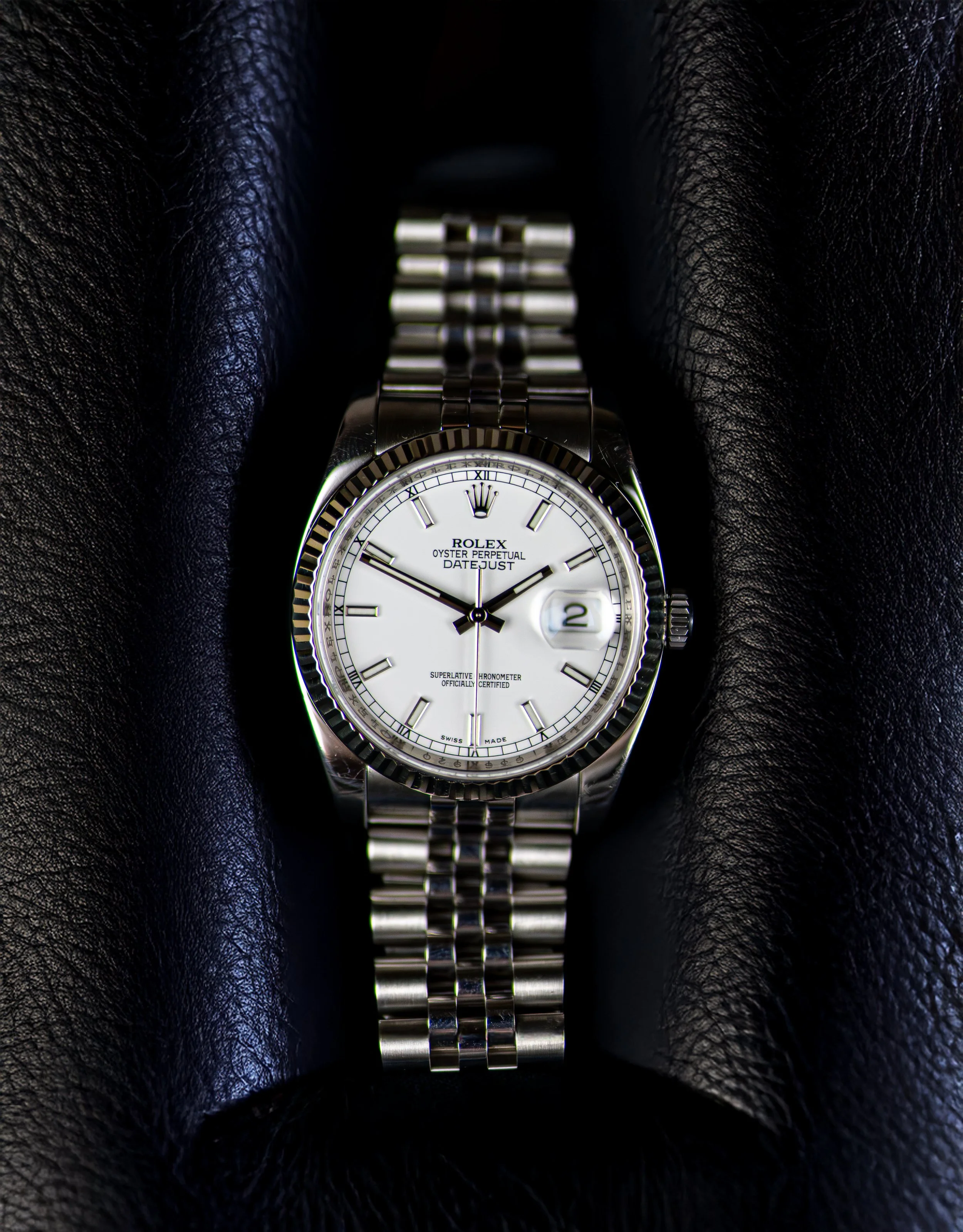 A silver Rolex Oyster Perpetual Datejust wristwatch with a white dial, Roman numeral hour markers, and a date display at 3 o'clock, resting inside black textured leather.