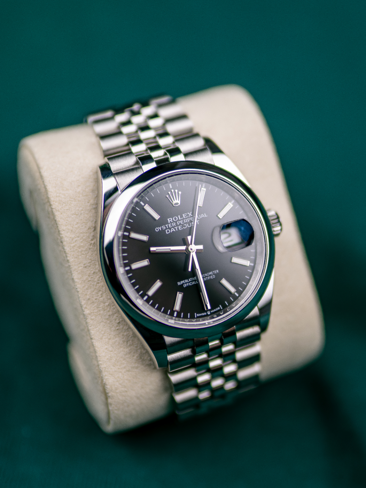 A close-up of a luxury Rolex Oyster Perpetual Datejust wristwatch with a black dial, silver indices, date display with magnifier, and a silver metal bracelet, resting on a beige cushion.
