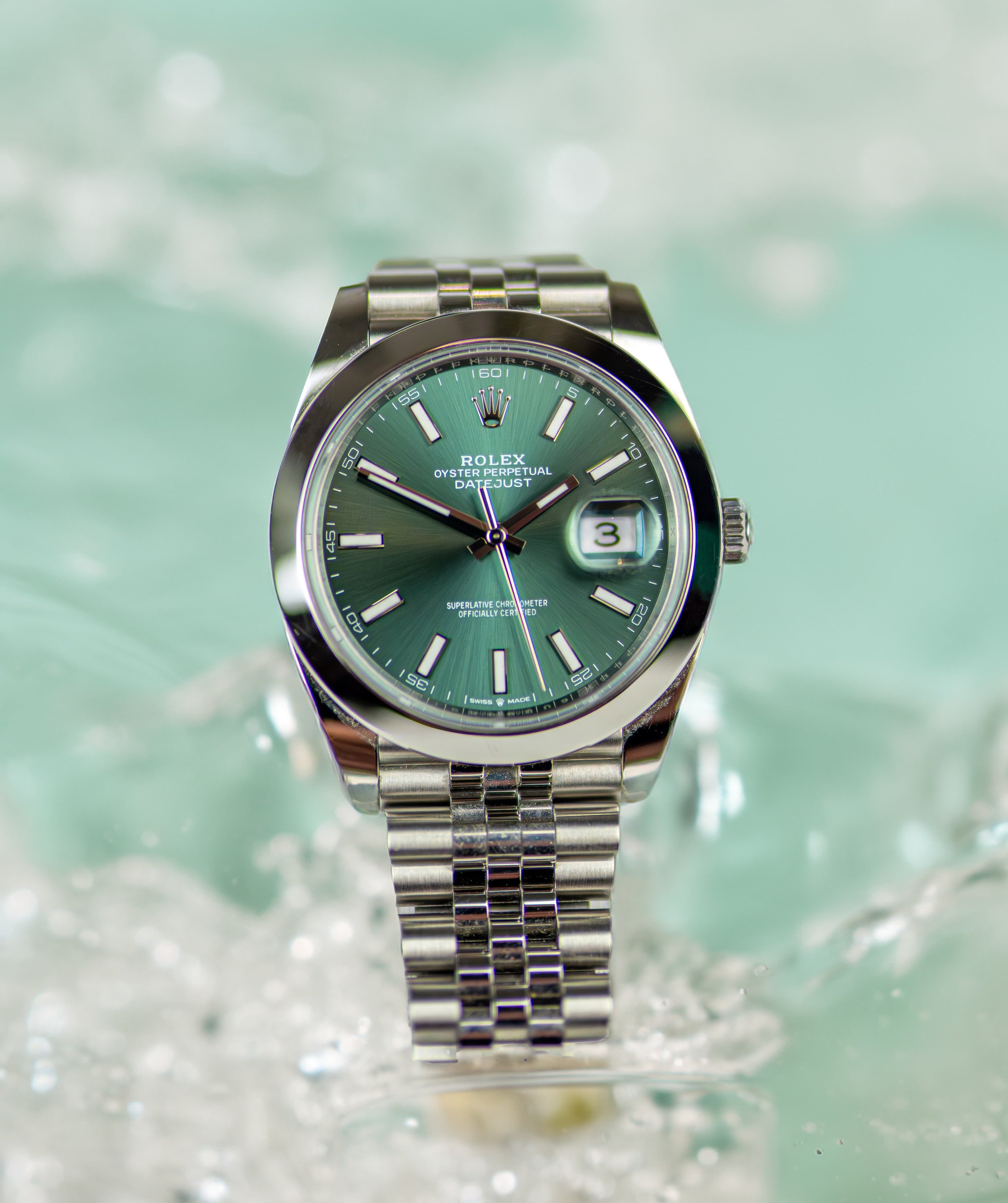 A green Rolex Oyster Perpetual Datejust watch with a stainless steel band, floating in water with bubbles and a soft, green background.