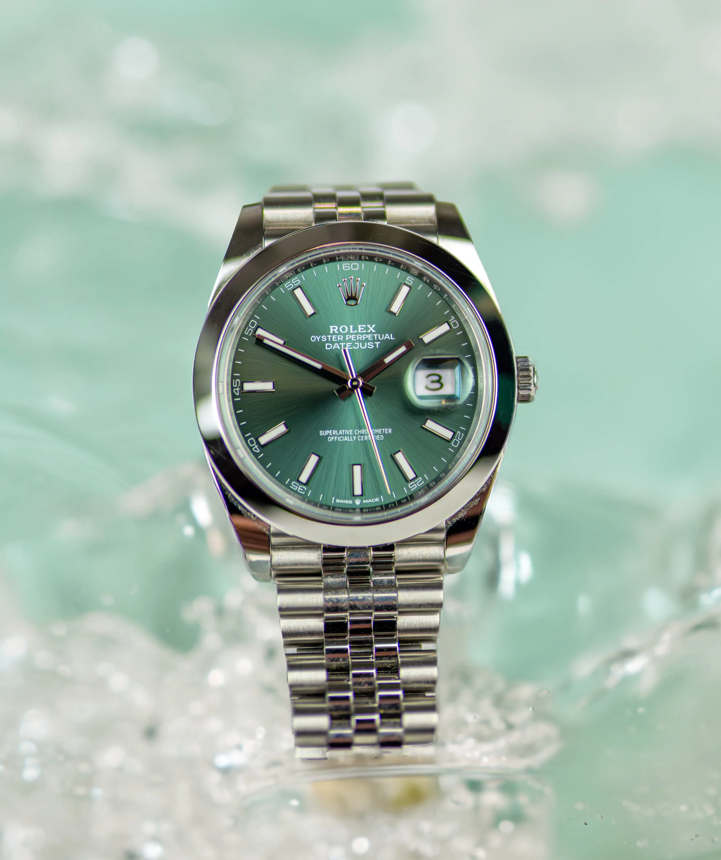 A silver Rolex Oyster Perpetual Datejust watch with a green dial, submerged in water with water splashes around it.