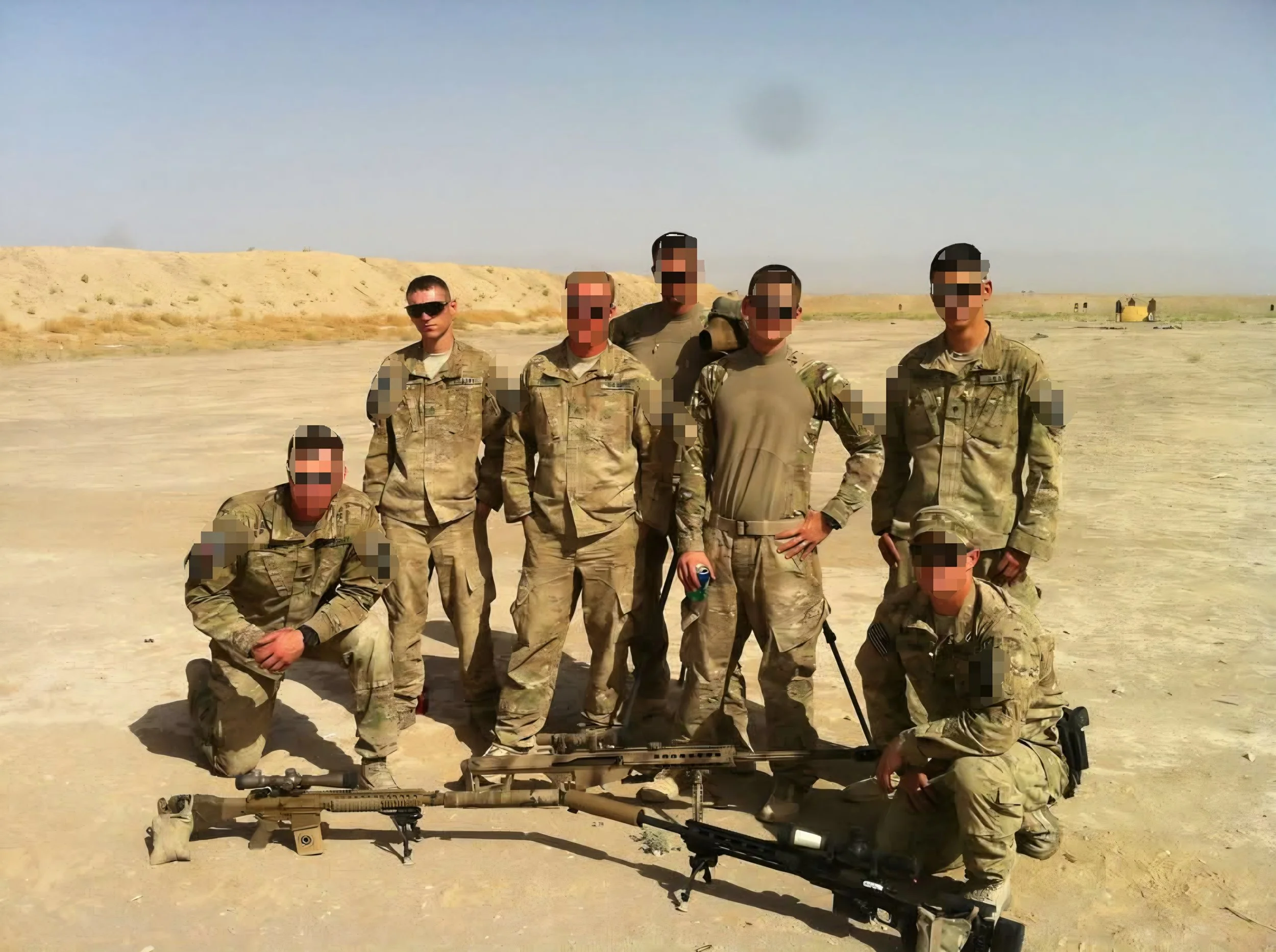 A group of five soldiers in desert camouflage uniforms standing outdoors on sandy terrain, with a clear blue sky, and some structures and smoke in the background.
