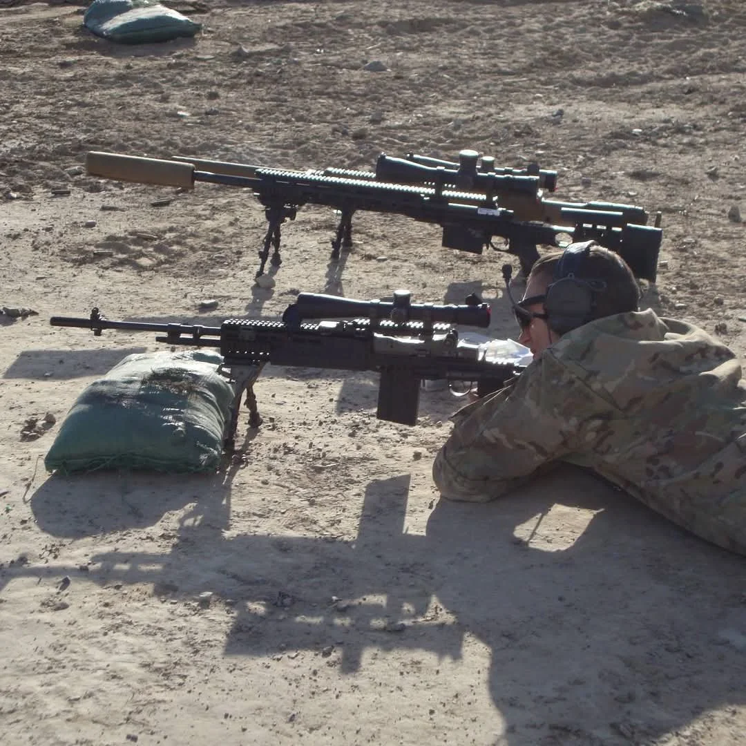 A soldier in camouflage uniform lying prone on the ground with two rifles aimed forward, one resting on a sandbag, on a dirt surface outside.