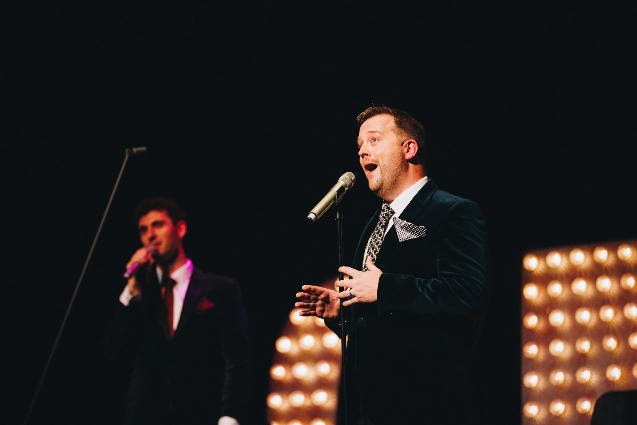 Two men in suits performing a song on stage, illuminated by warm stage lights.