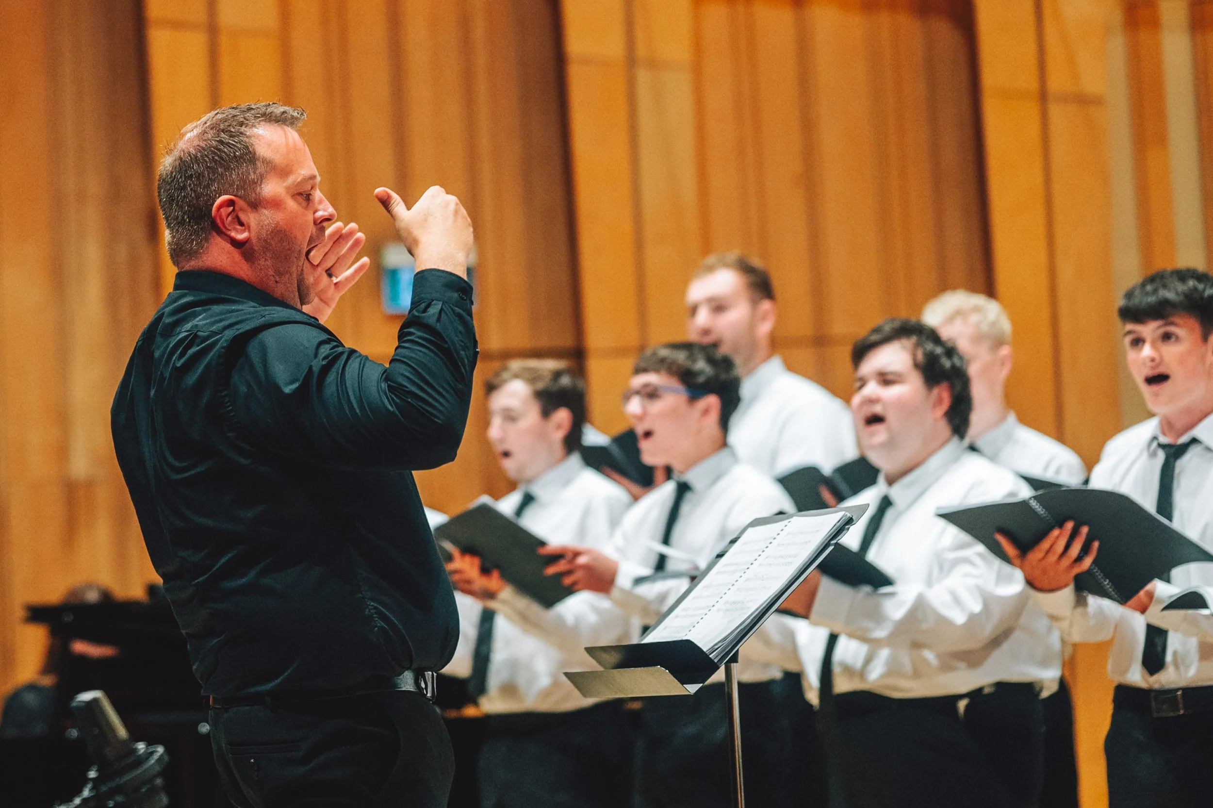 Craig leading a group of young male singers during a rehearsal in a wooden-paneled room.