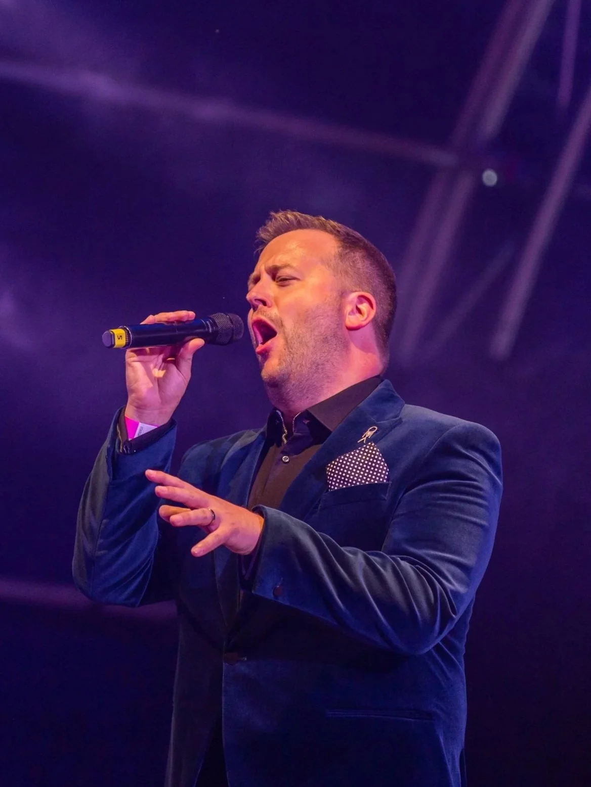 Craig Yates singing into a microphone on stage, wearing a dark suit with a polka dot pocket square.