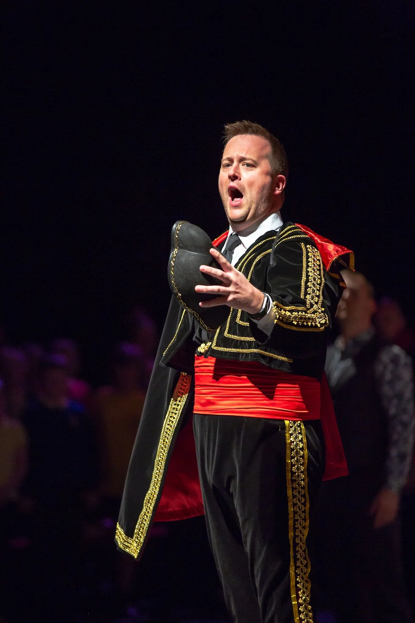 Craig Yates dressed in a traditional bullfighter costume, standing on a stage, holding his hat, with a serious expression, in front of a dark background with a few out-of-focus people.