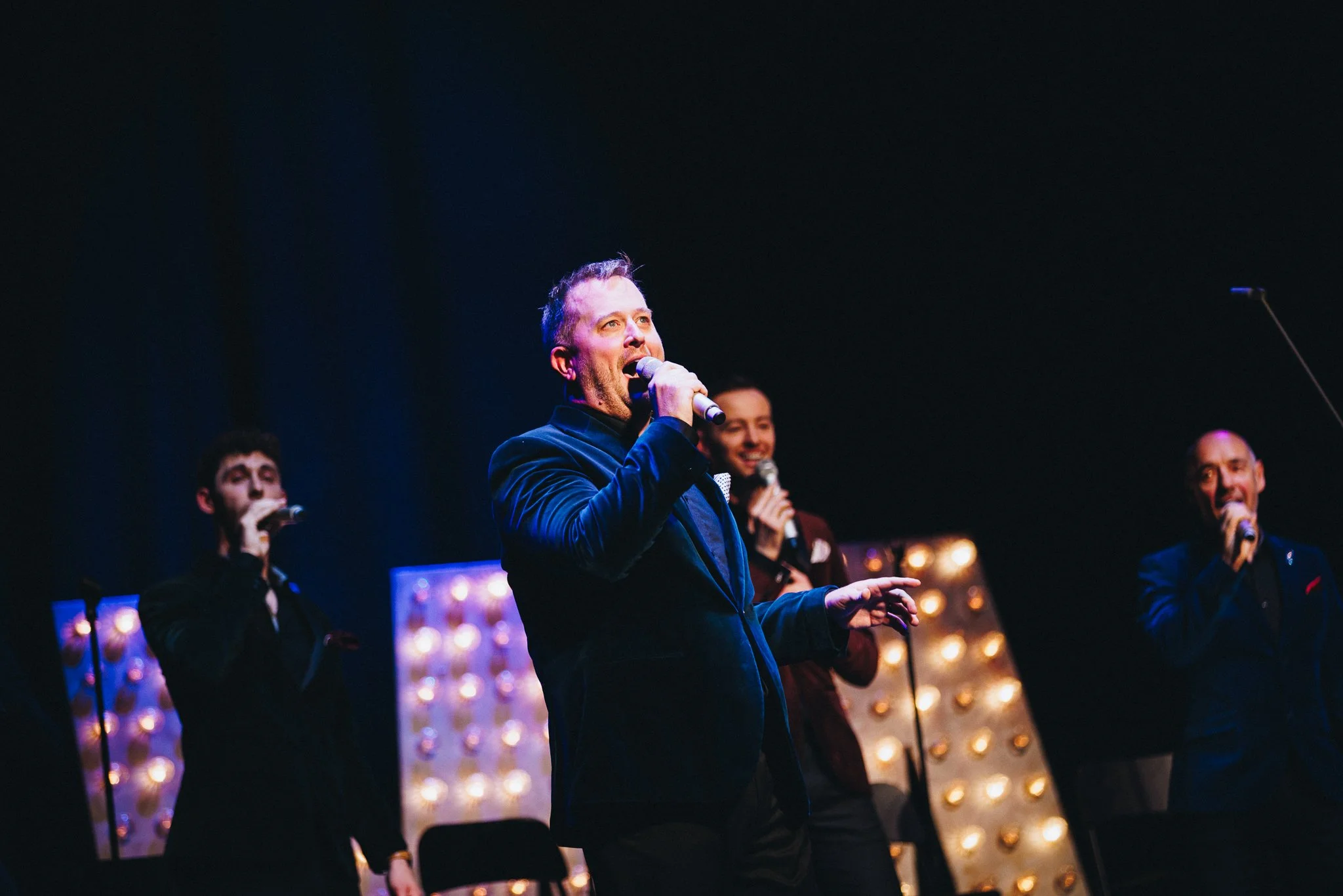 Four men performing with Only Men Aloud on stage with microphones, illuminated decorative lights in the background, and a dark overall setting.