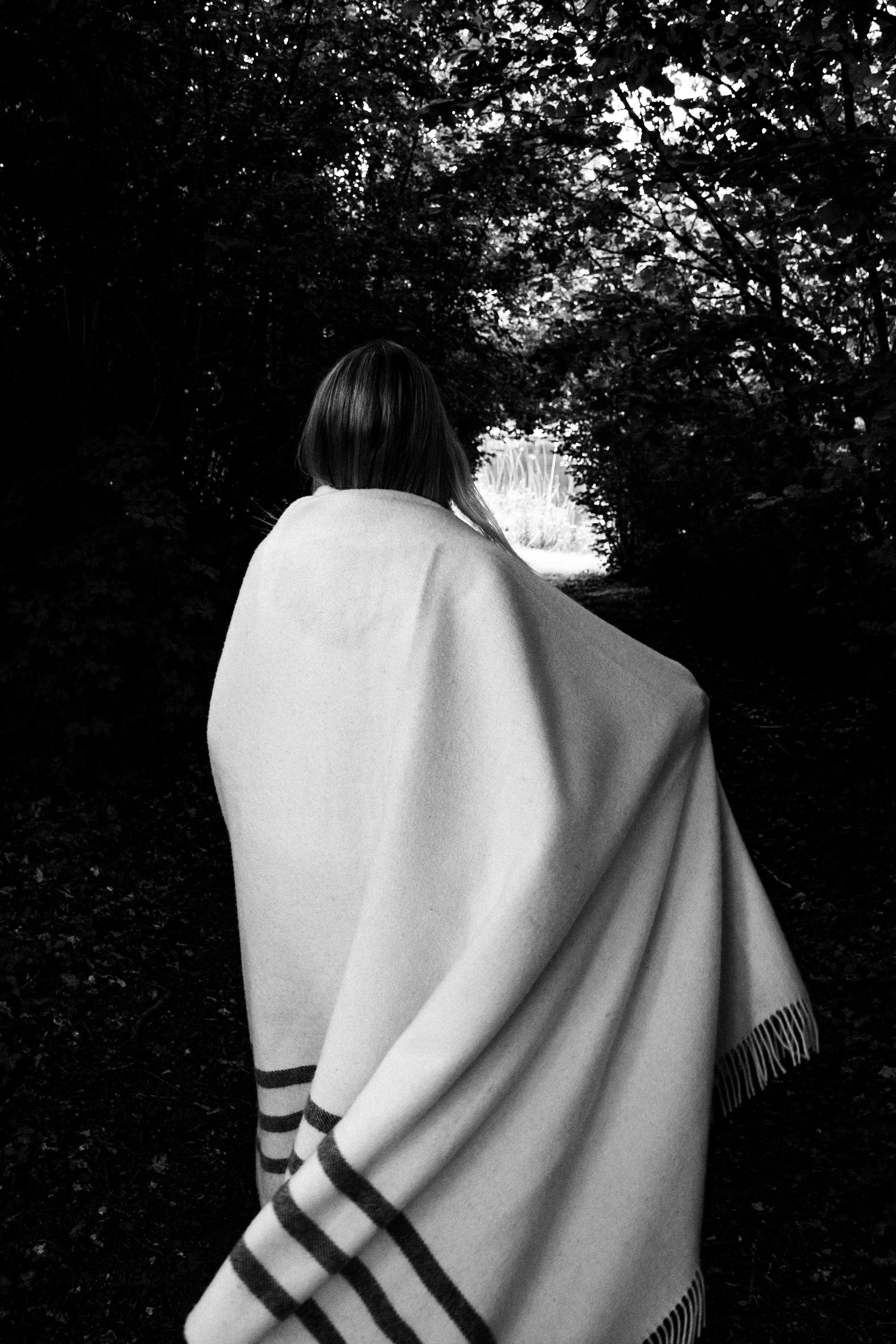 A woman with straight hair seen from behind, wrapped in a large blanket with striped edges, walking through a dark, tree-lined tunnel towards a bright opening.