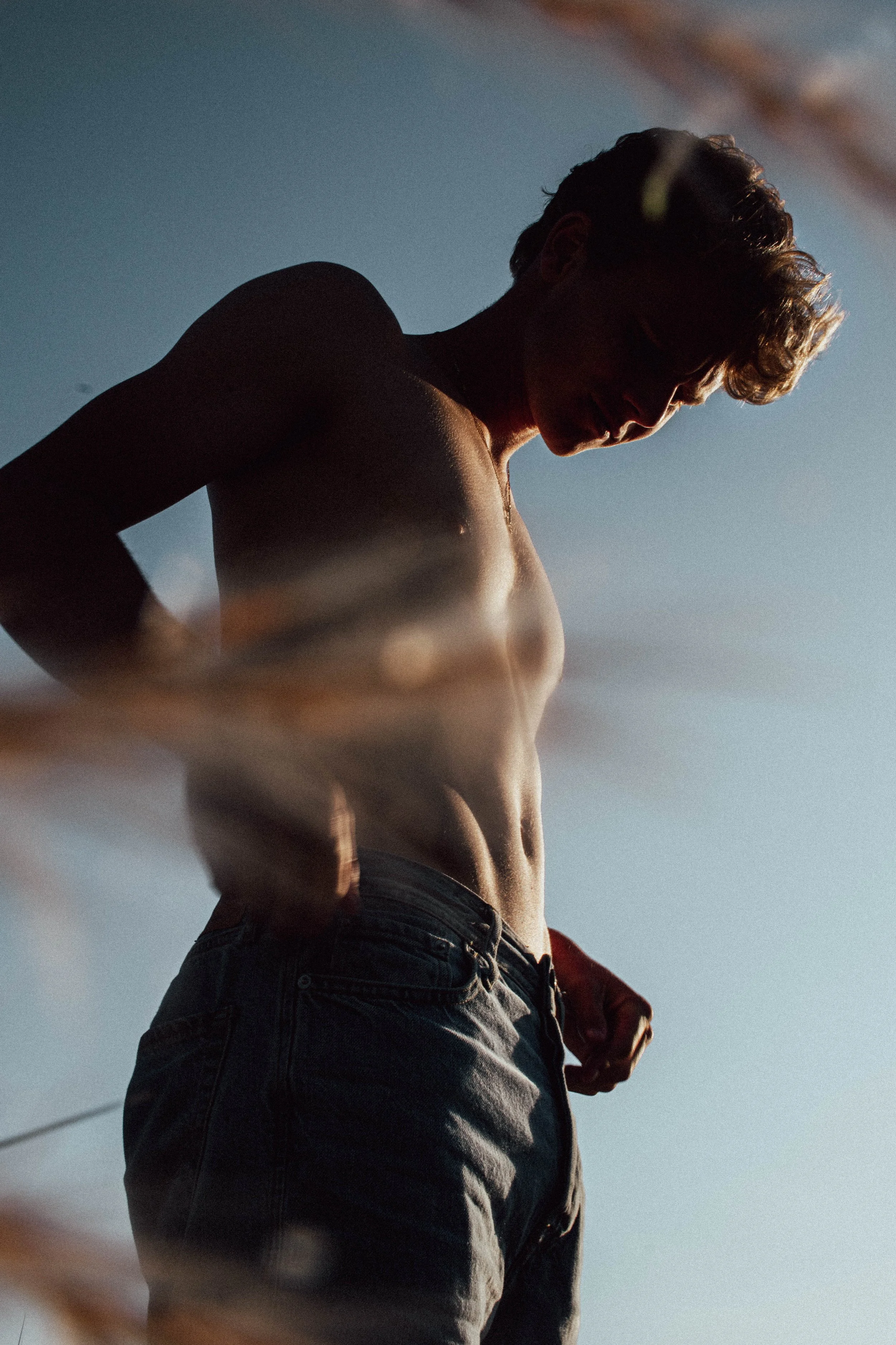 A shirtless young man with curly hair looking down, standing outdoors against a clear sky, with sunlight highlighting his torso and face.