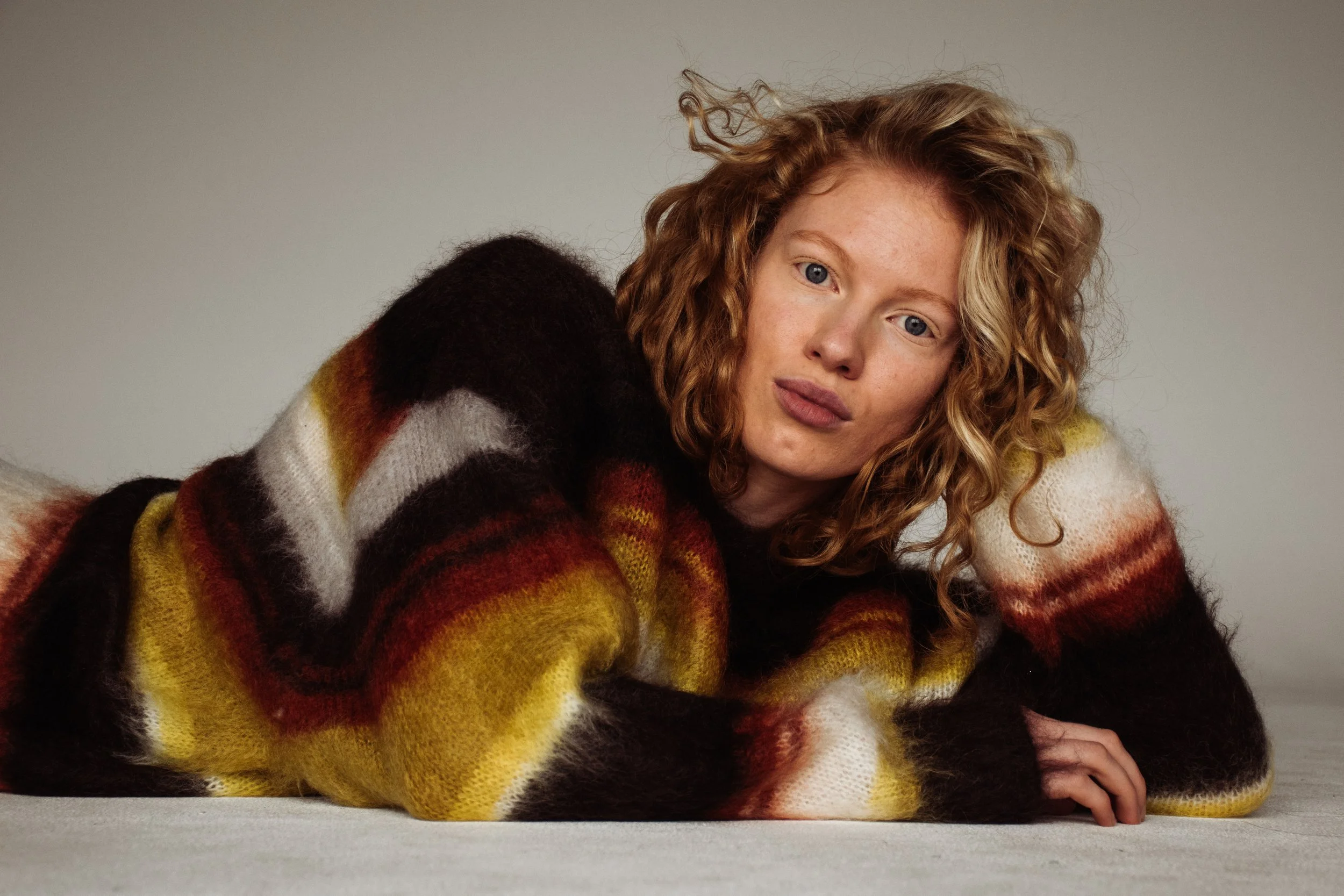 A woman with curly red hair lying on a flat surface, wearing a multicolored, striped, fuzzy sweater, with a neutral background.