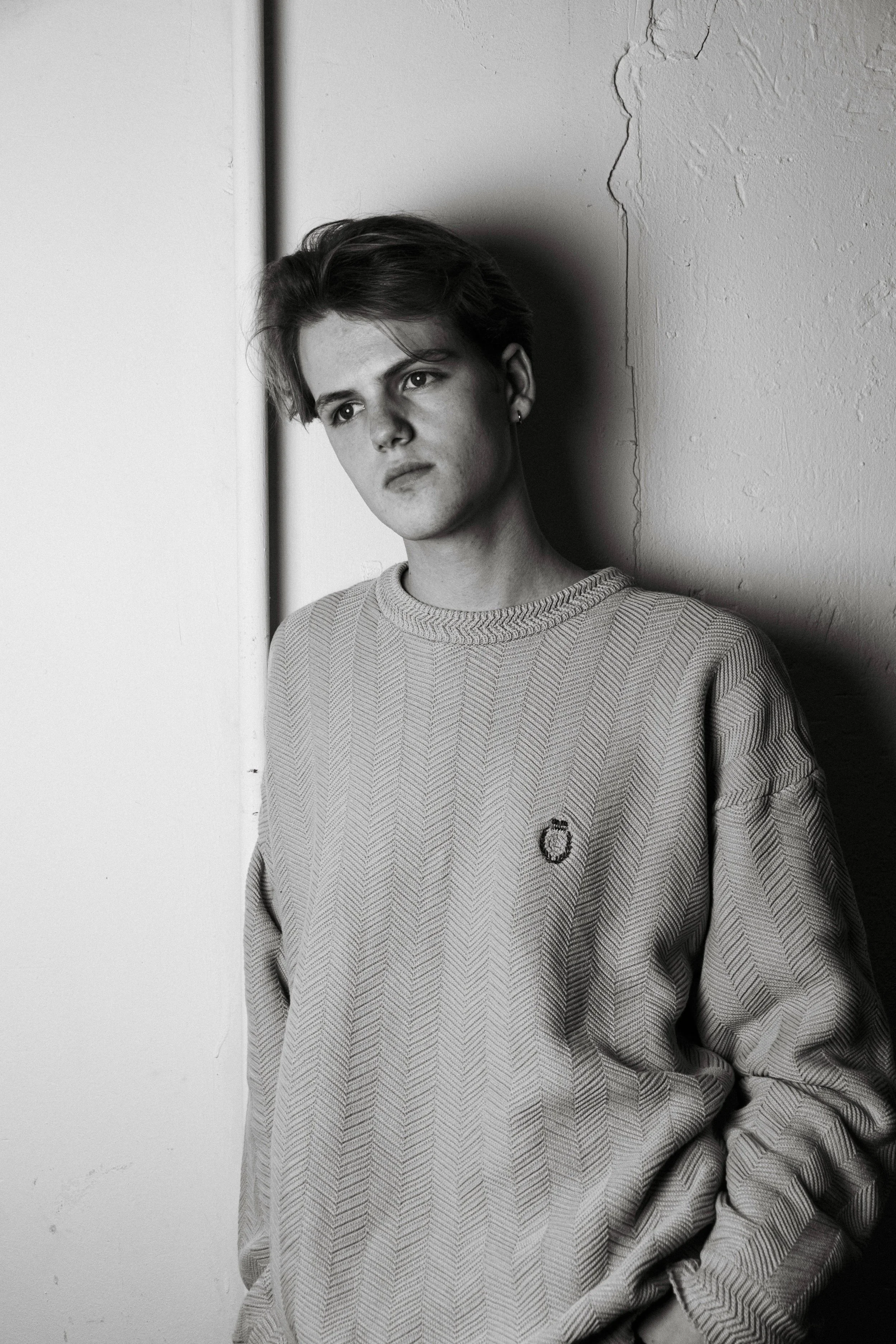 A young person with short hair leaning against a wall, staring thoughtfully.