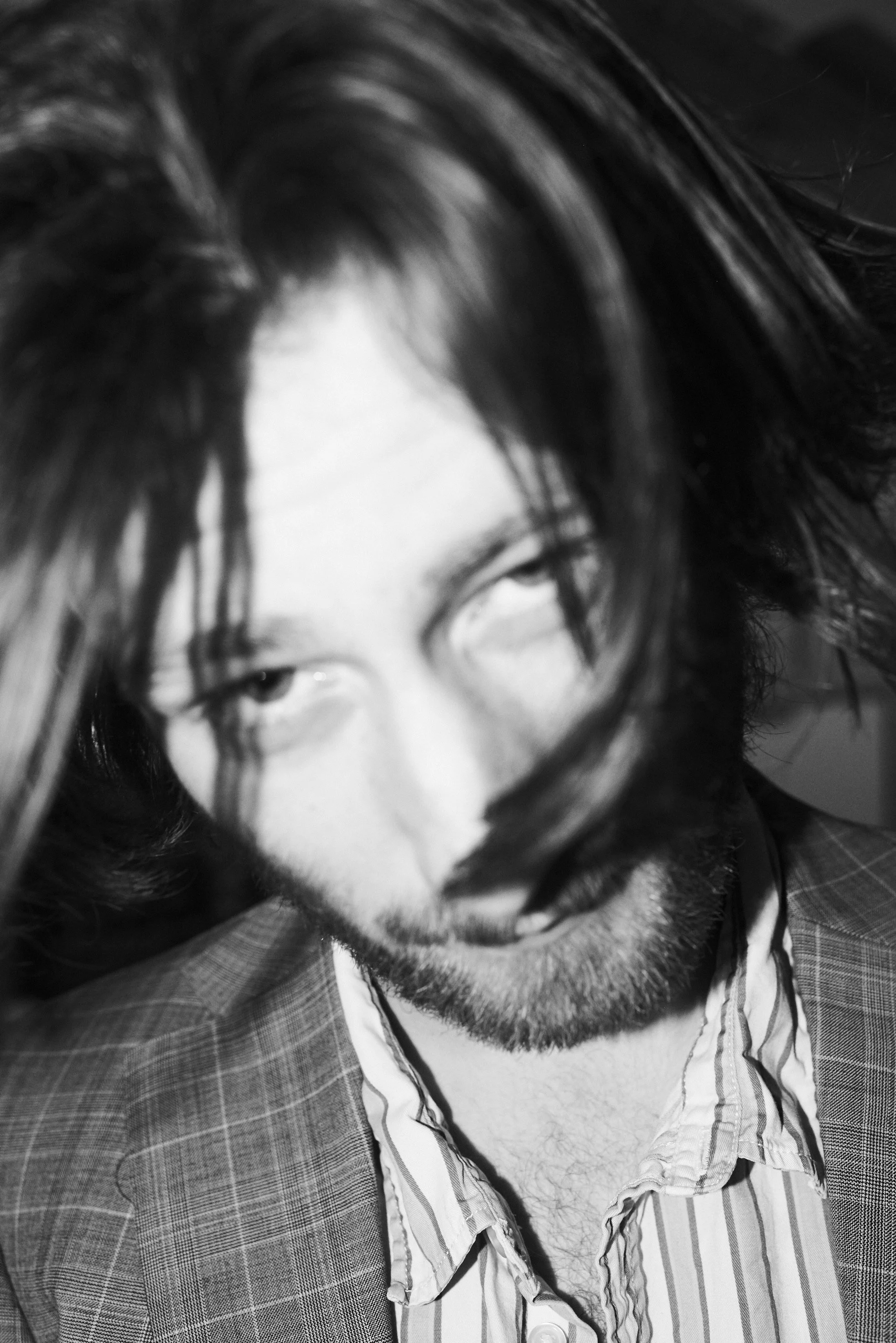 A black and white close-up photograph of a man with long hair, a beard, and wearing a checkered blazer and a striped shirt, looking directly at the camera with a serious expression.