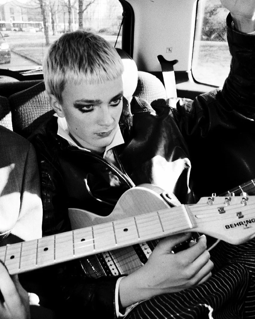 A young person with short hair and dramatic eye makeup sitting in a vehicle, holding an electric guitar, with someone else partially visible playing another guitar.