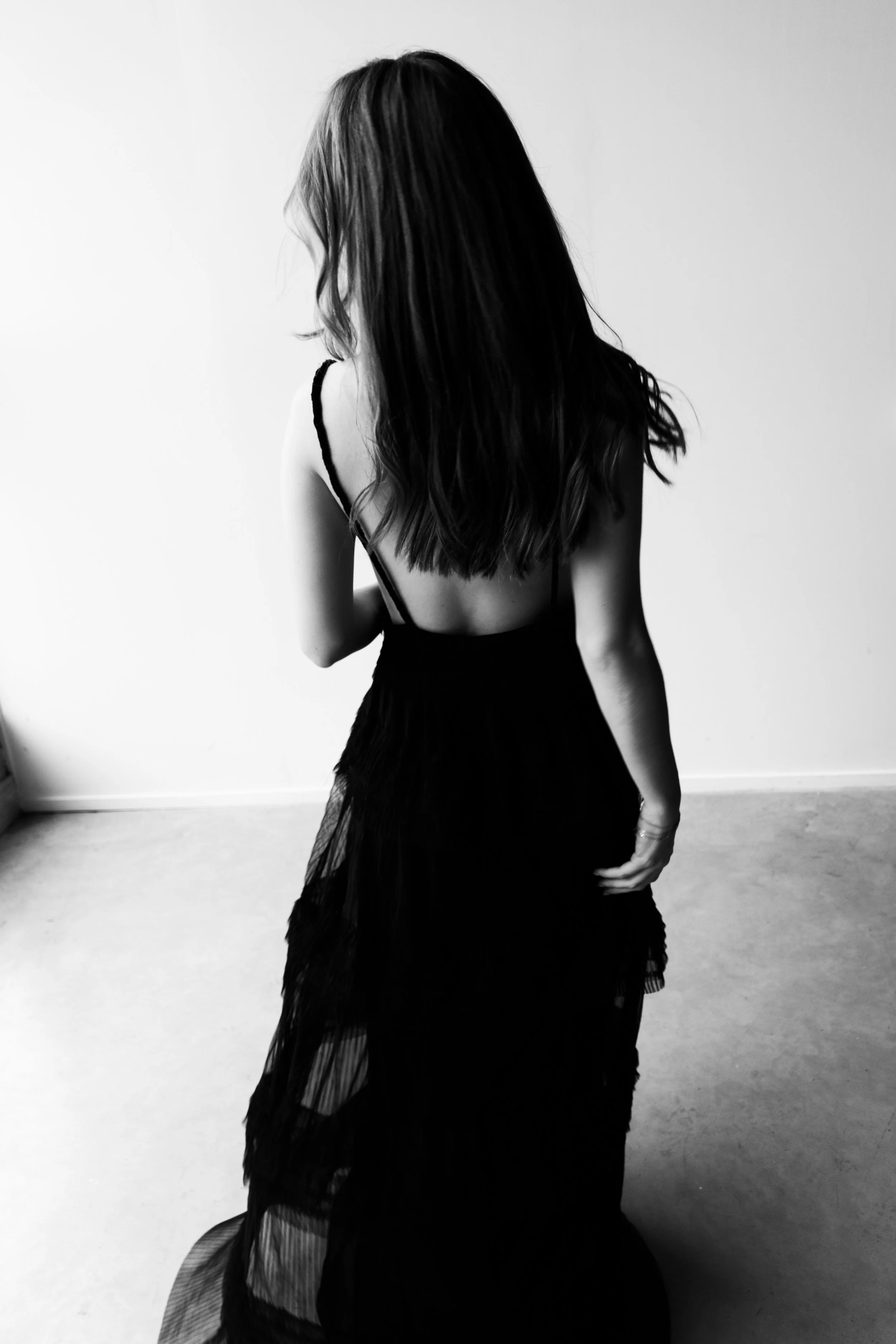 Black and white photo of a woman with long hair, wearing a backless dress, standing indoors against a plain wall.