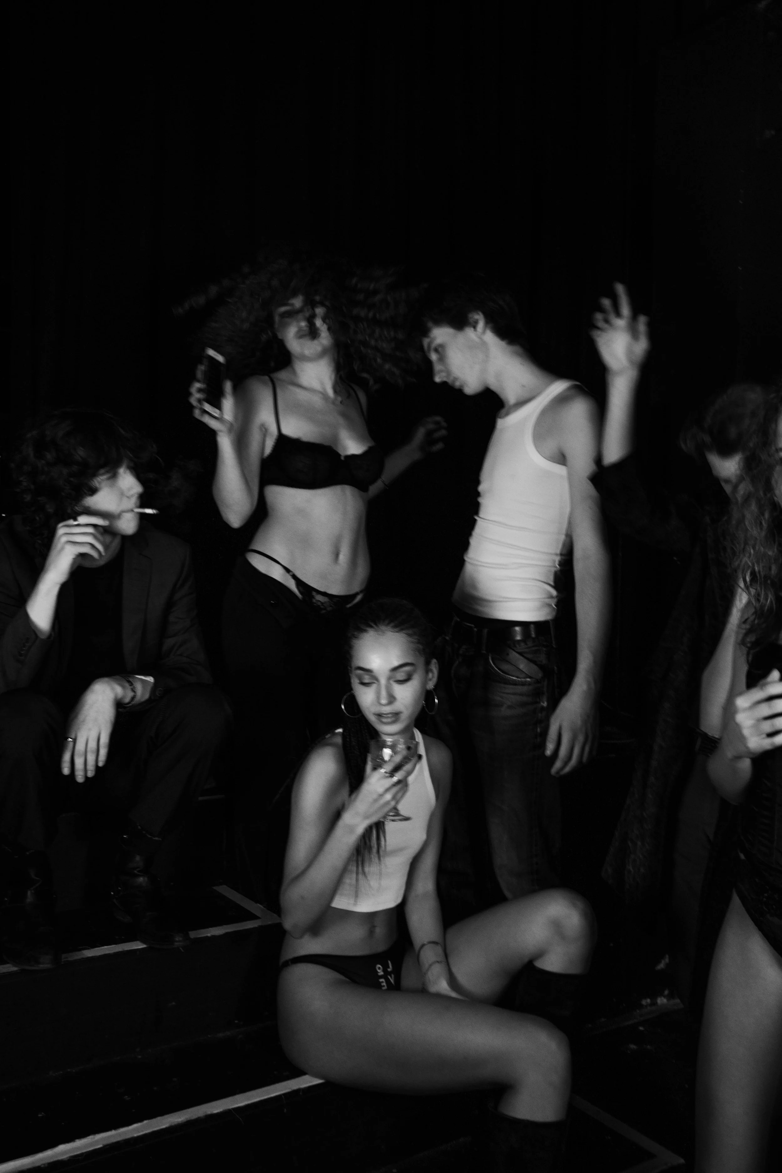 Black and white photo of five young adults at a party. One woman is sitting on the floor holding a drink, while the others are standing or sitting nearby. Two are smoking, and one is looking at their phone, with a dark backdrop behind them.