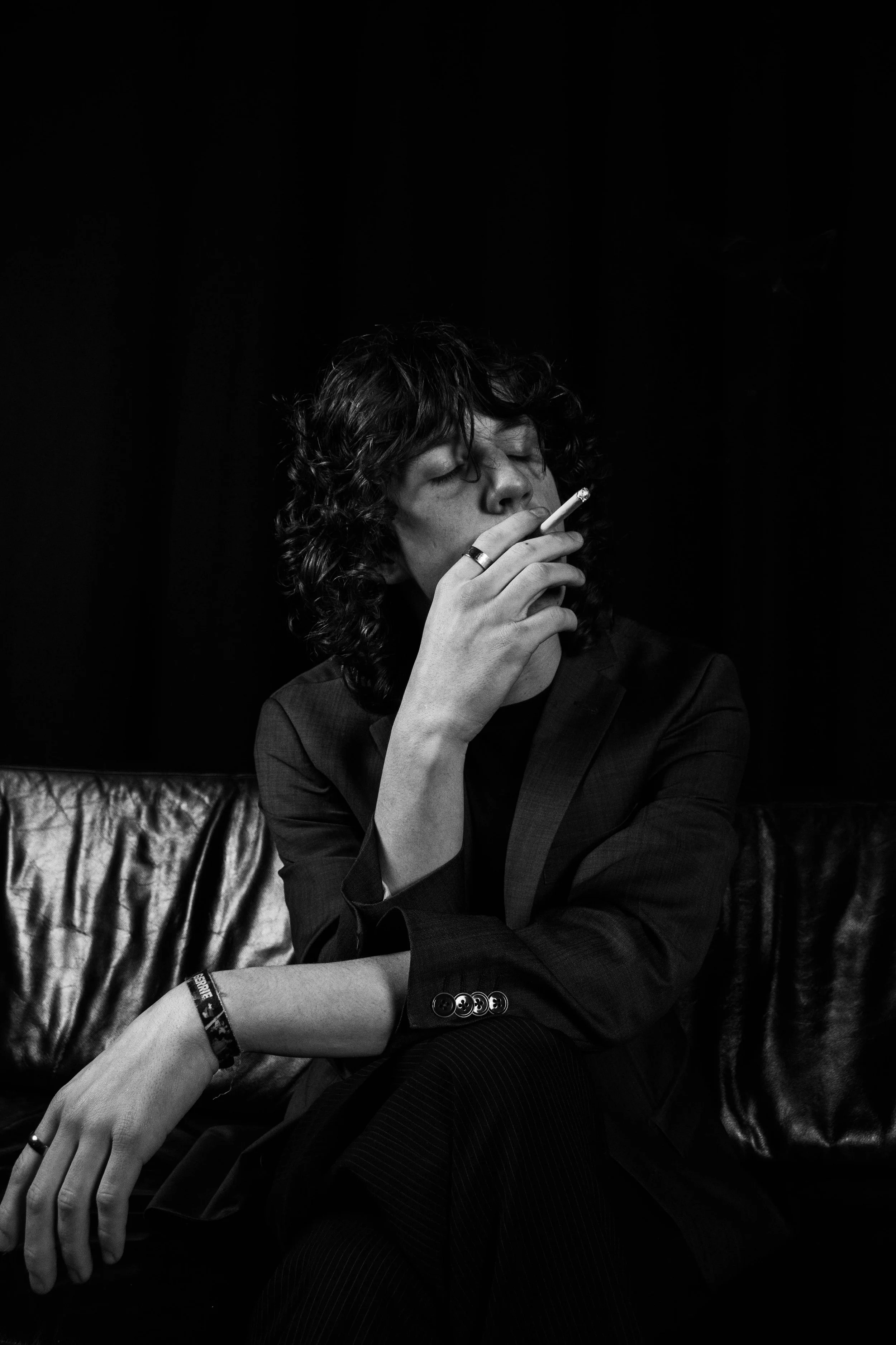 A black and white photo of a person with curly hair, wearing a blazer, sitting on a shiny leather couch, smoking a cigarette with eyes closed.