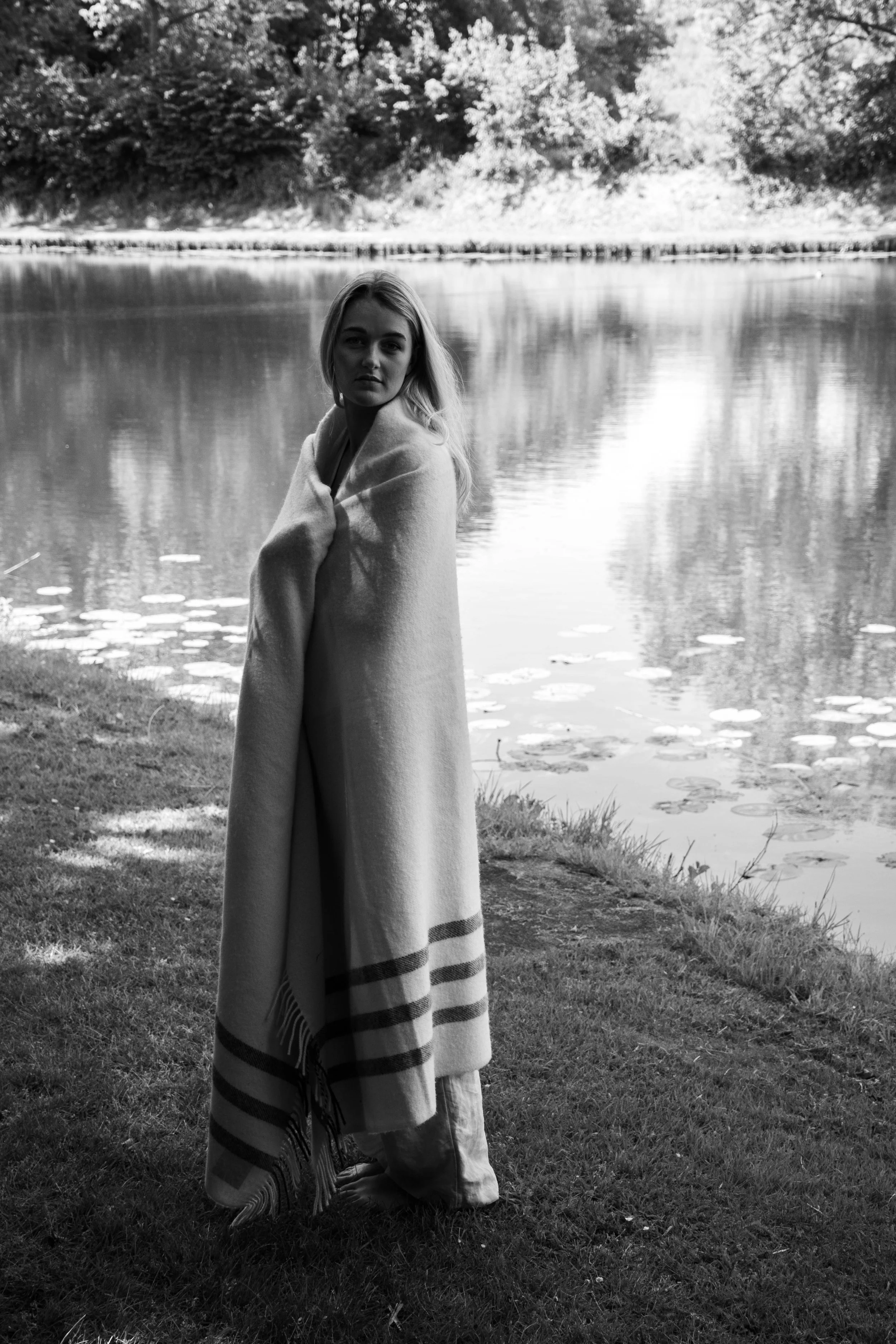 A woman standing on grass near a lake, wrapped in a large blanket with striped edges, in black and white.