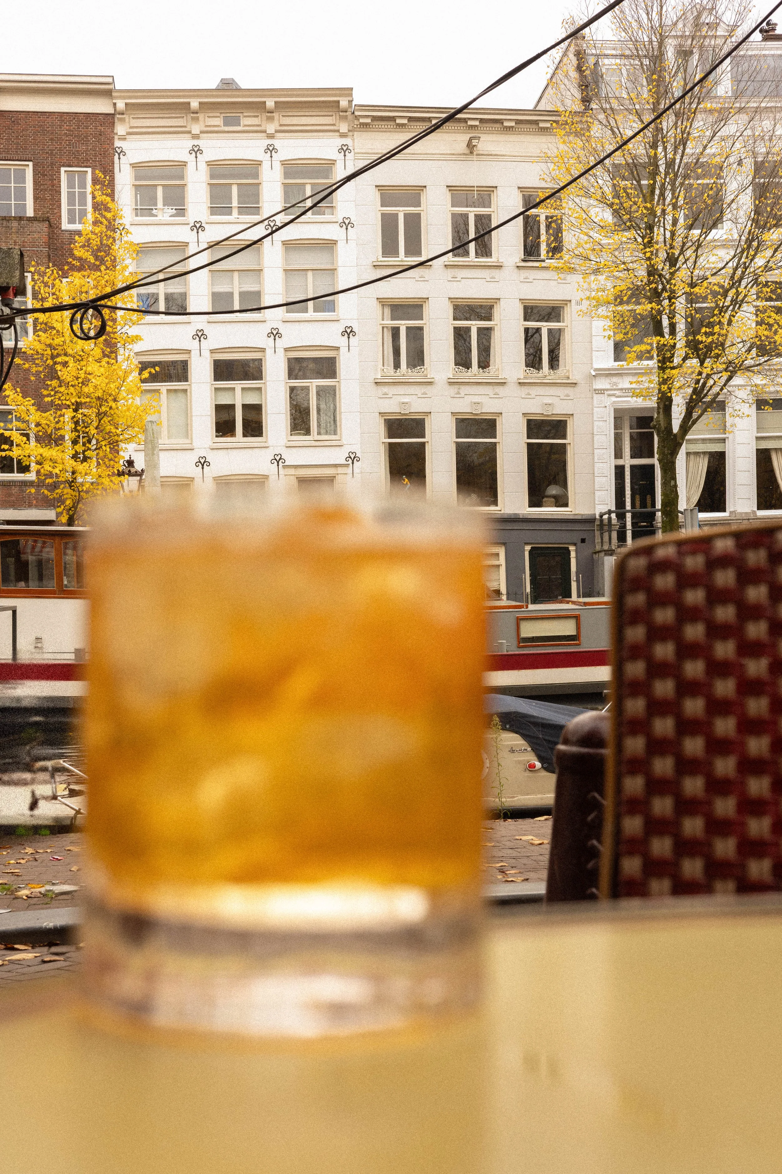 A blurry glass of yellow-colored drink on a table with a city street view of buildings, trees with autumn leaves, and a canal with boats in the background.