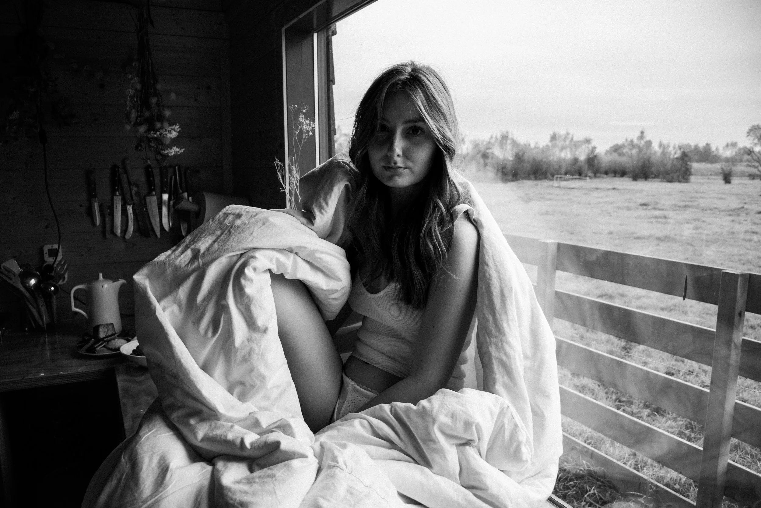 A woman with long hair sitting on a bed near a large window, with a countryside landscape visible outside. The room has knives hanging on the wall and various objects on a wooden table.