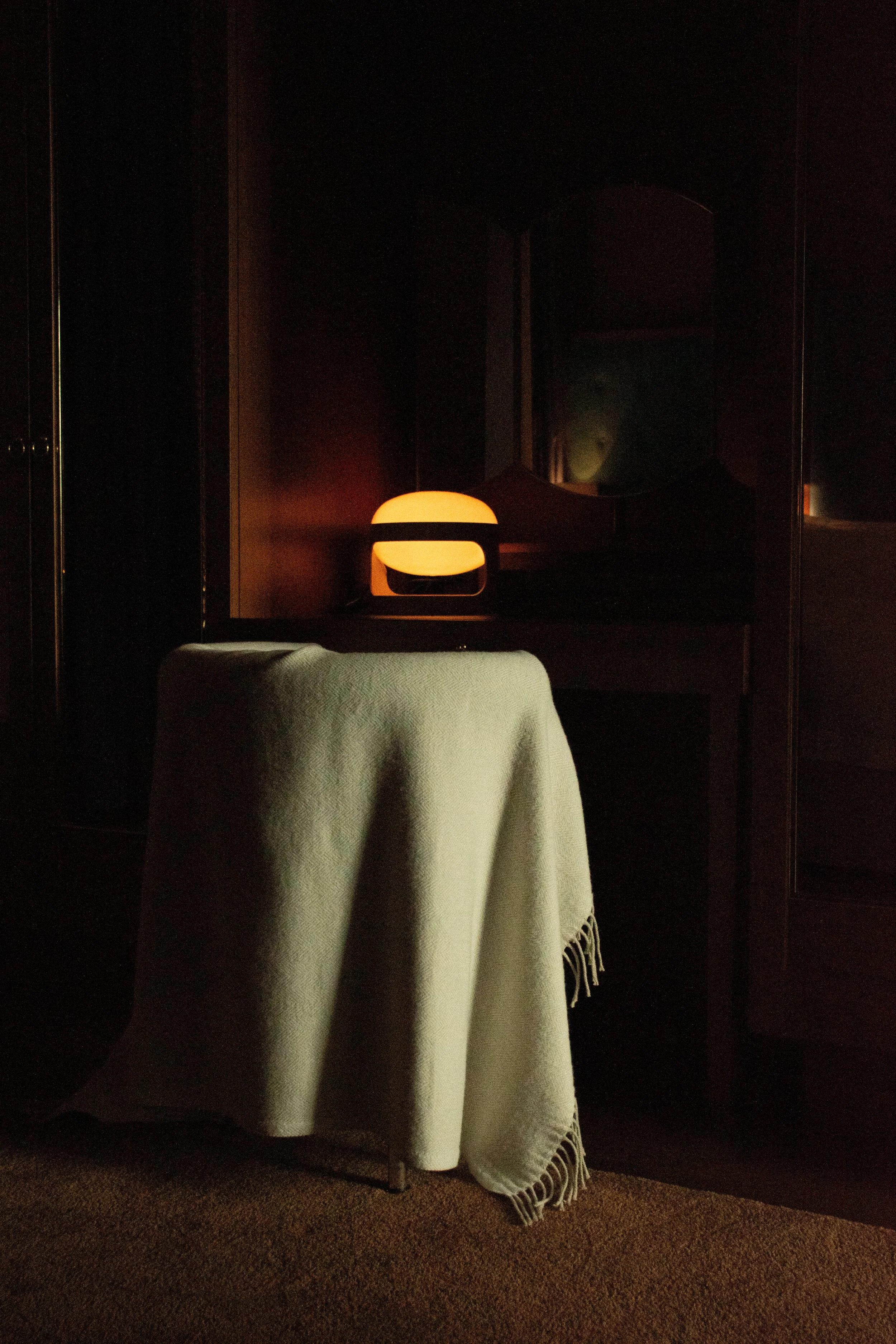 A dimly lit room with a beige blanket draped over a chair, a small lamp emitting warm light, and a mirror on the dark wooden wall behind.