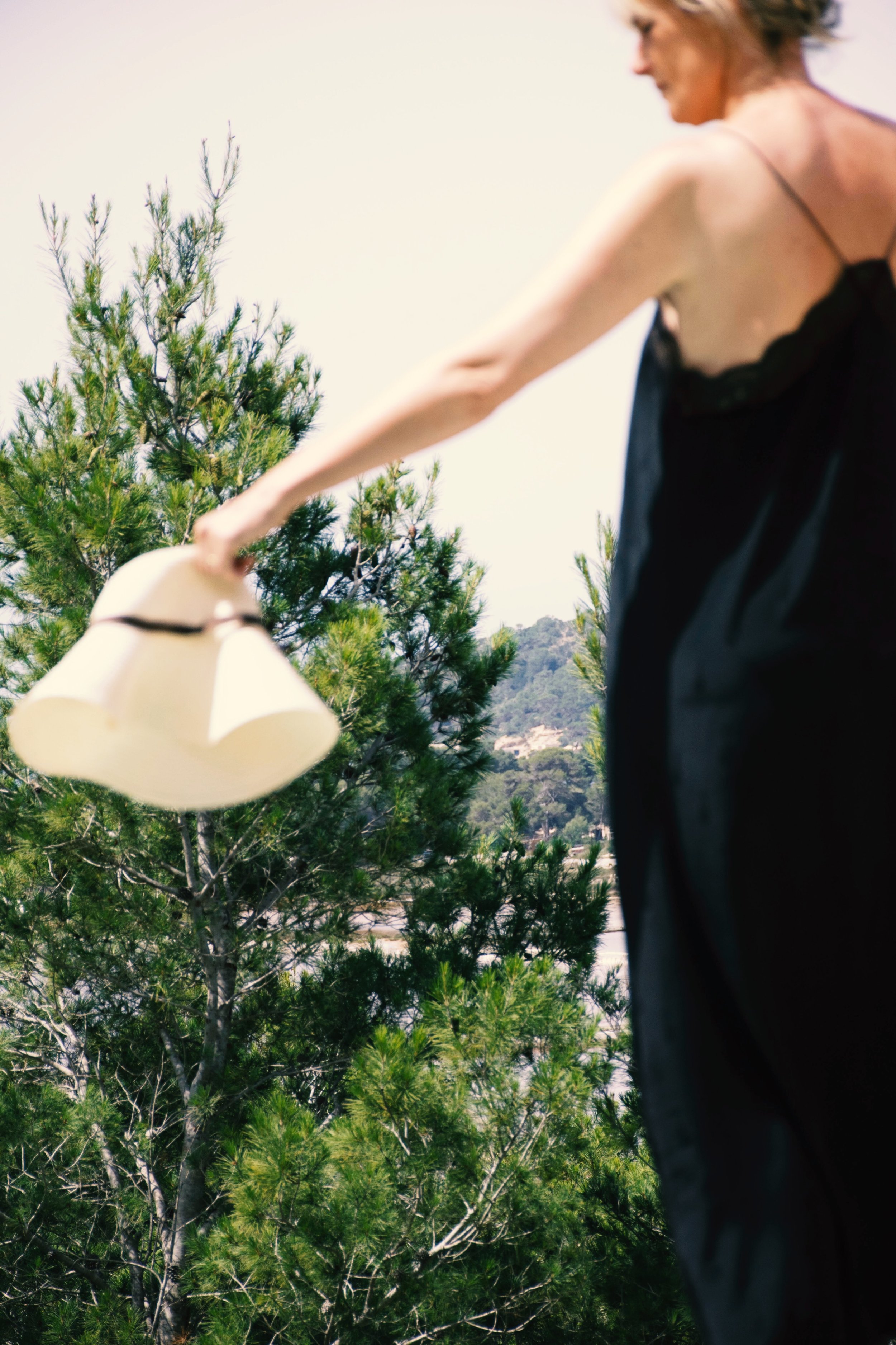 A woman in a black dress holding a white hat outdoors with green trees and a distant landscape in the background.