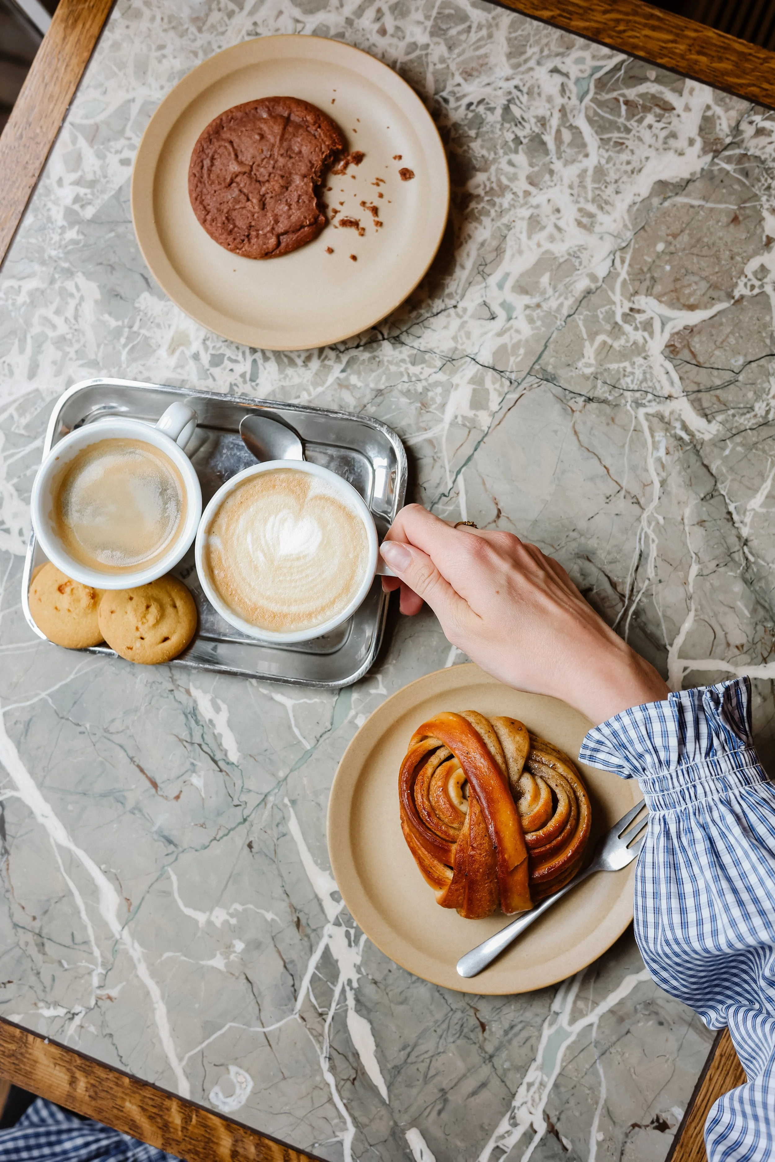 A breakfast scene on a marble table showing a plate with a cinnamon roll, two cups of coffee with foam art, a small tray with a spoon and two cookies, and a person's hand holding the tray with a cinnamon bun and fork.