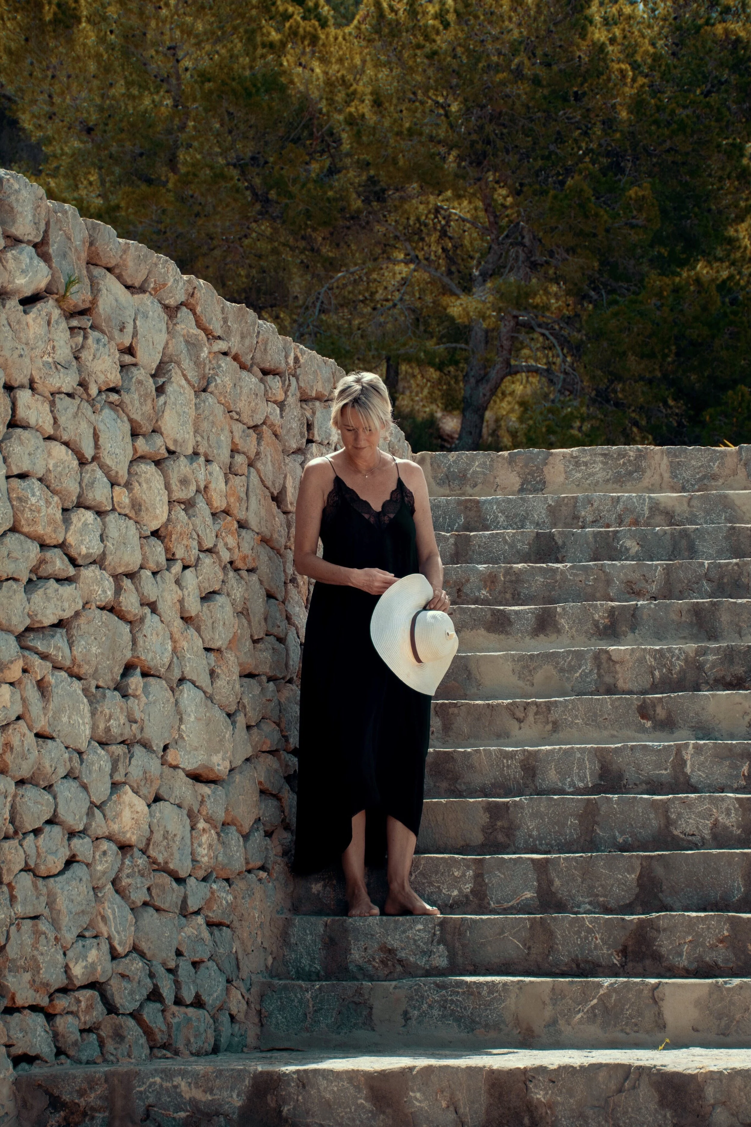 A woman in a black dress holding a straw hat standing on stone steps next to a stone wall, with trees in the background.