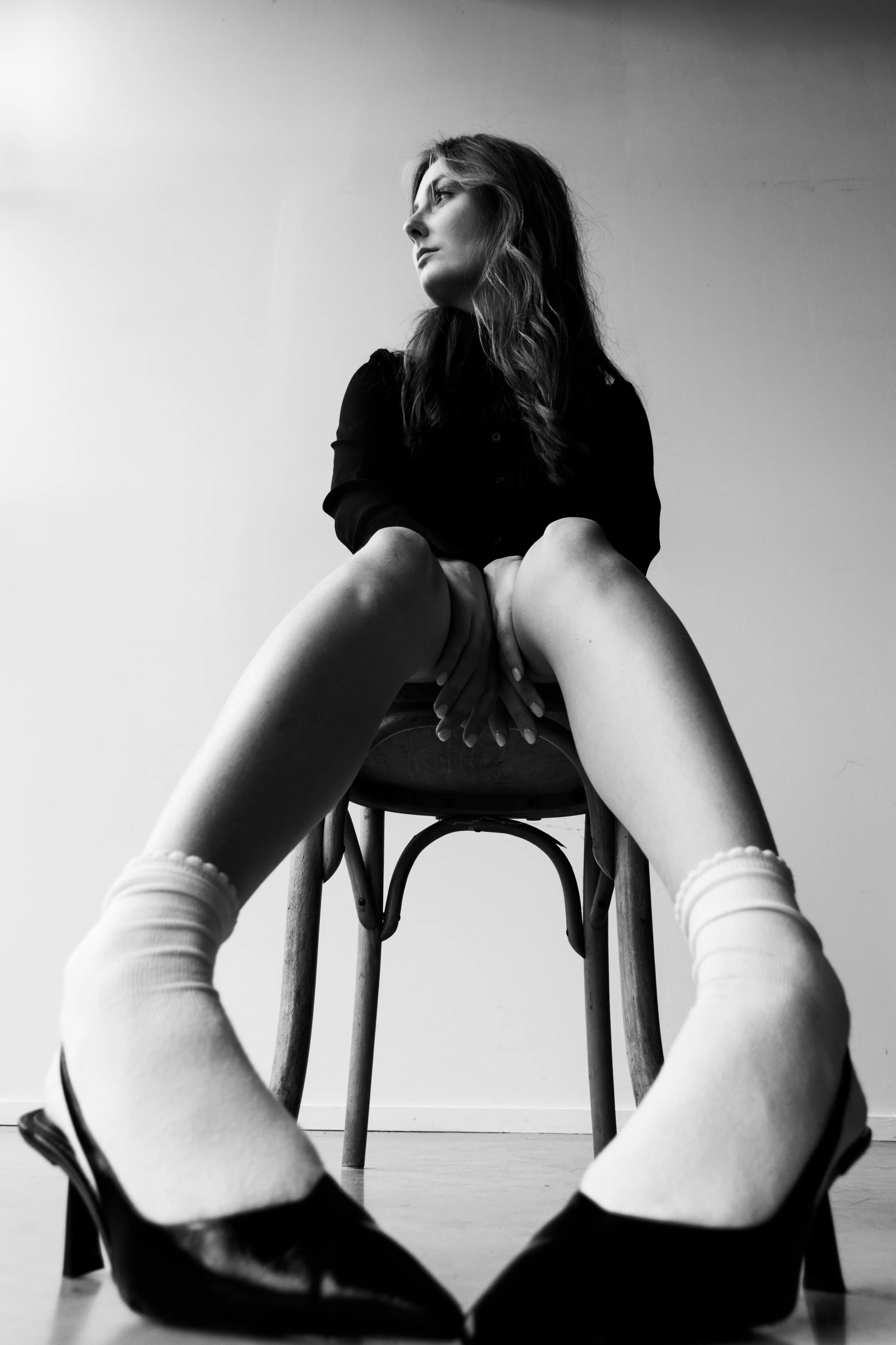 Black and white photo of a woman sitting on a chair with her legs apart, wearing high heels and white socks, looking to her left with her hands resting on her lap.