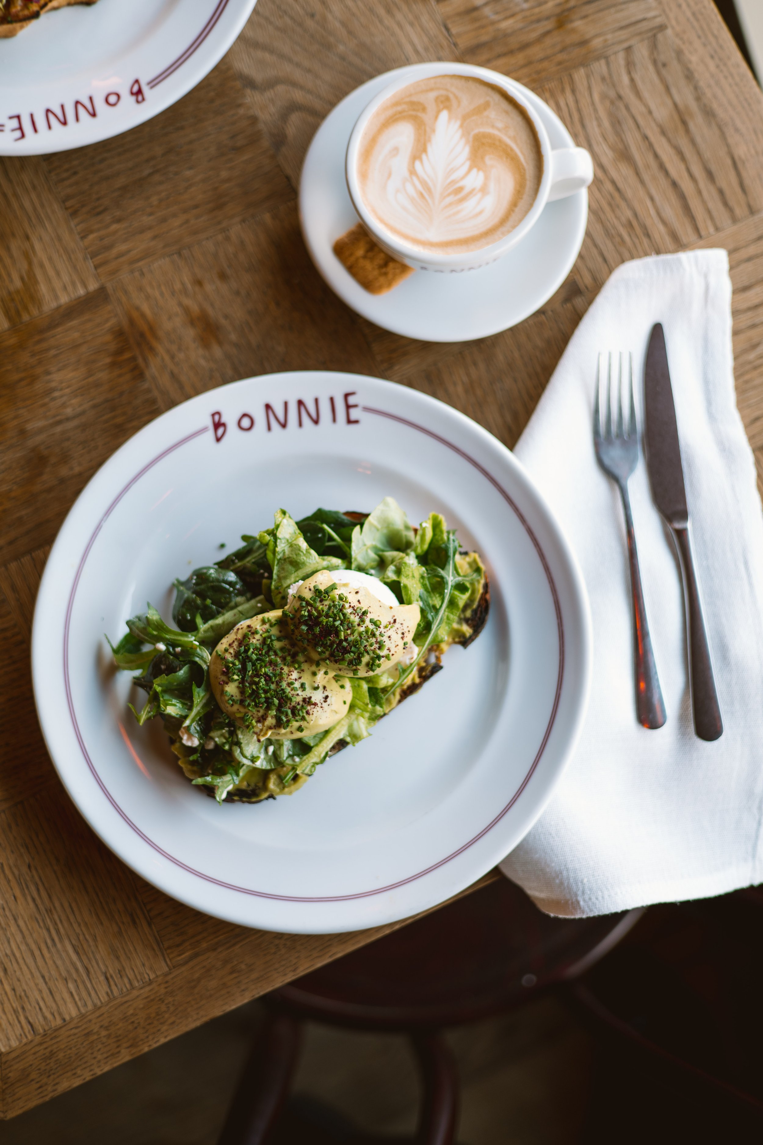 A breakfast setting with a plate of eggs Benedict topped with chopped chives, a side of mixed greens, a cup of latte with latte art, a biscuit, a fork, and a knife on a white napkin, all on a wooden table.