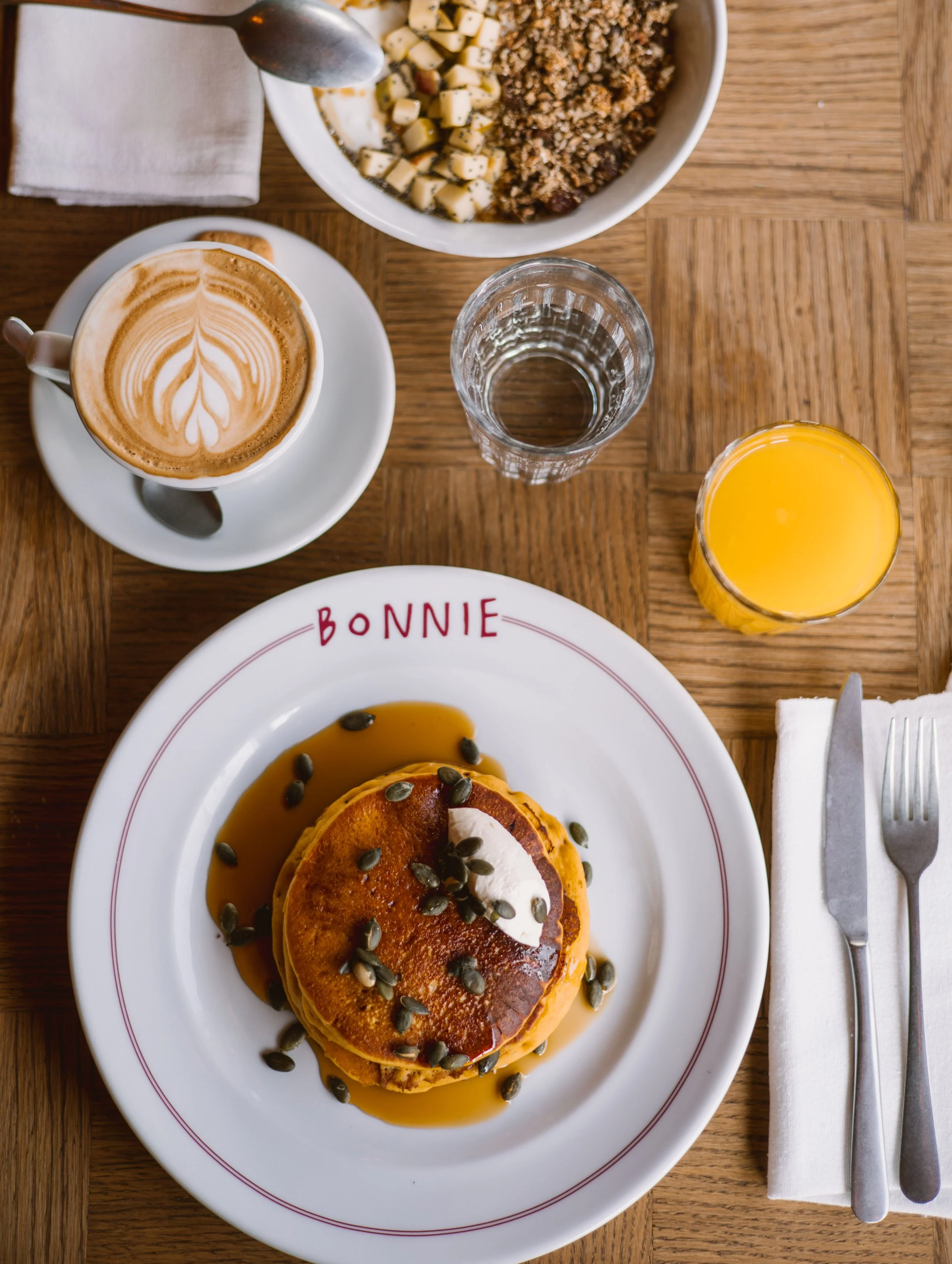 Breakfast table with a plate of pancakes topped with whipped cream and pumpkin seeds, a cup of latte with latte art, a glass of orange juice, a glass of water, and a bowl of cereal with milk.