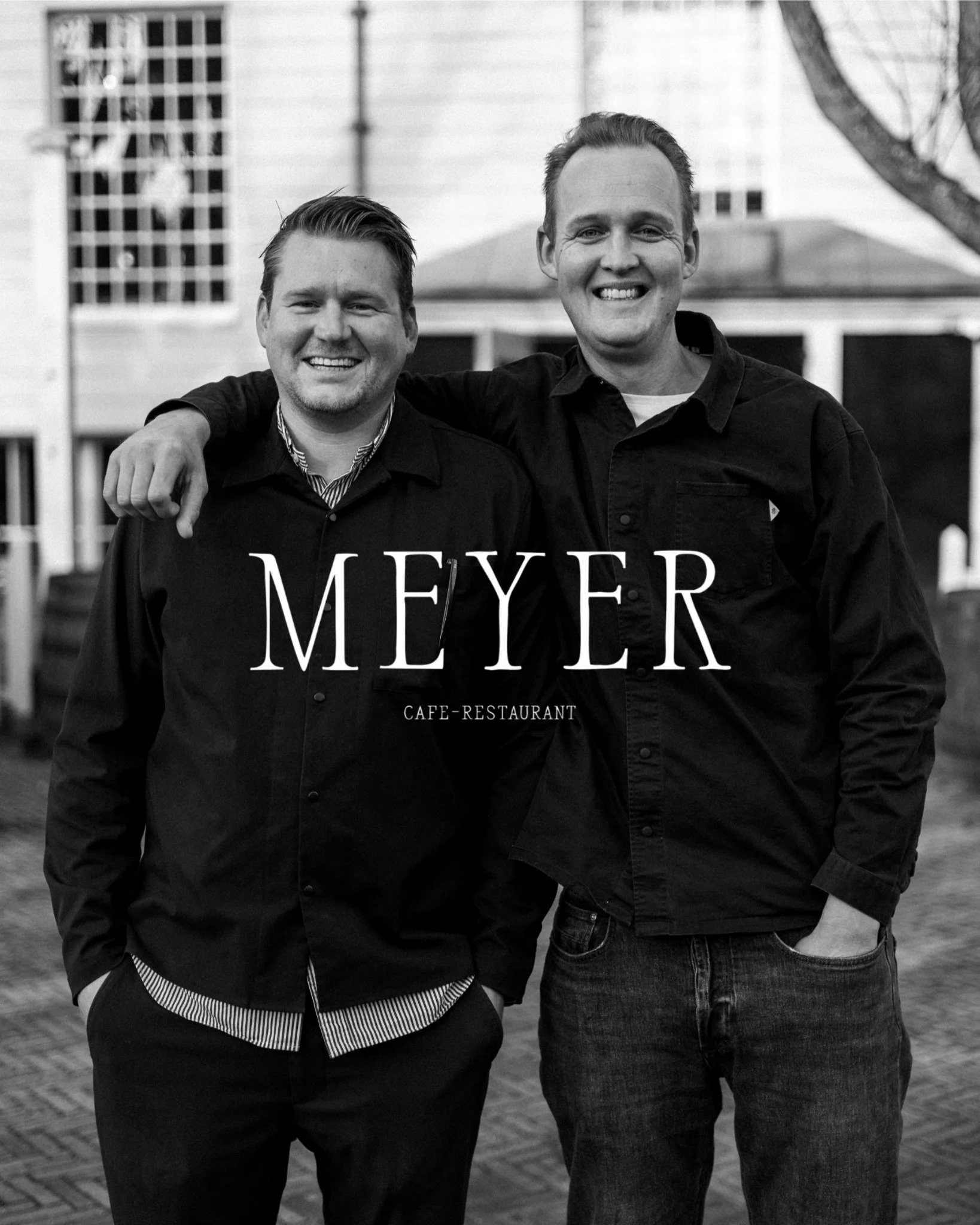 Two men smiling and standing side by side outdoors, with one man resting his arm on the other’s shoulder. They are wearing dark shirts, and there are buildings and a tree in the background. The text 'Meyer Cafe-Restaurant' is overlaid on the image.