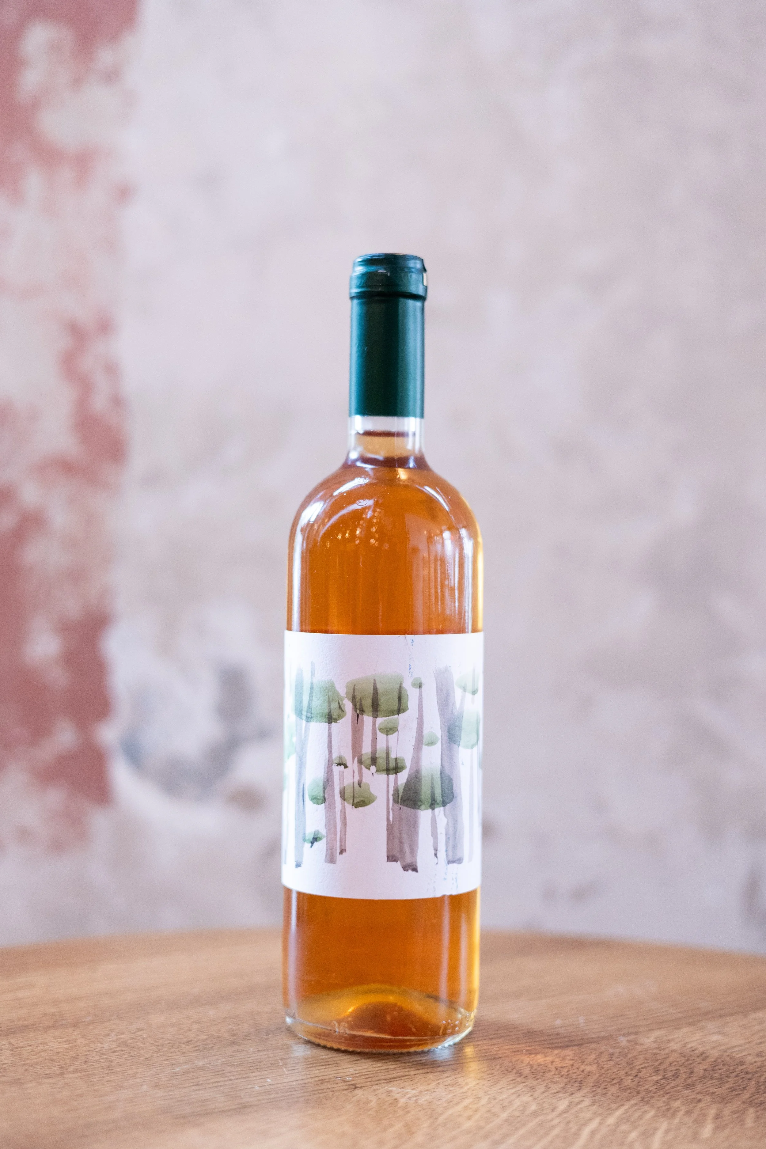 A bottle of rosé wine with a green cap and a white label featuring abstract watercolor trees, placed on a wooden surface with a textured wall in the background.