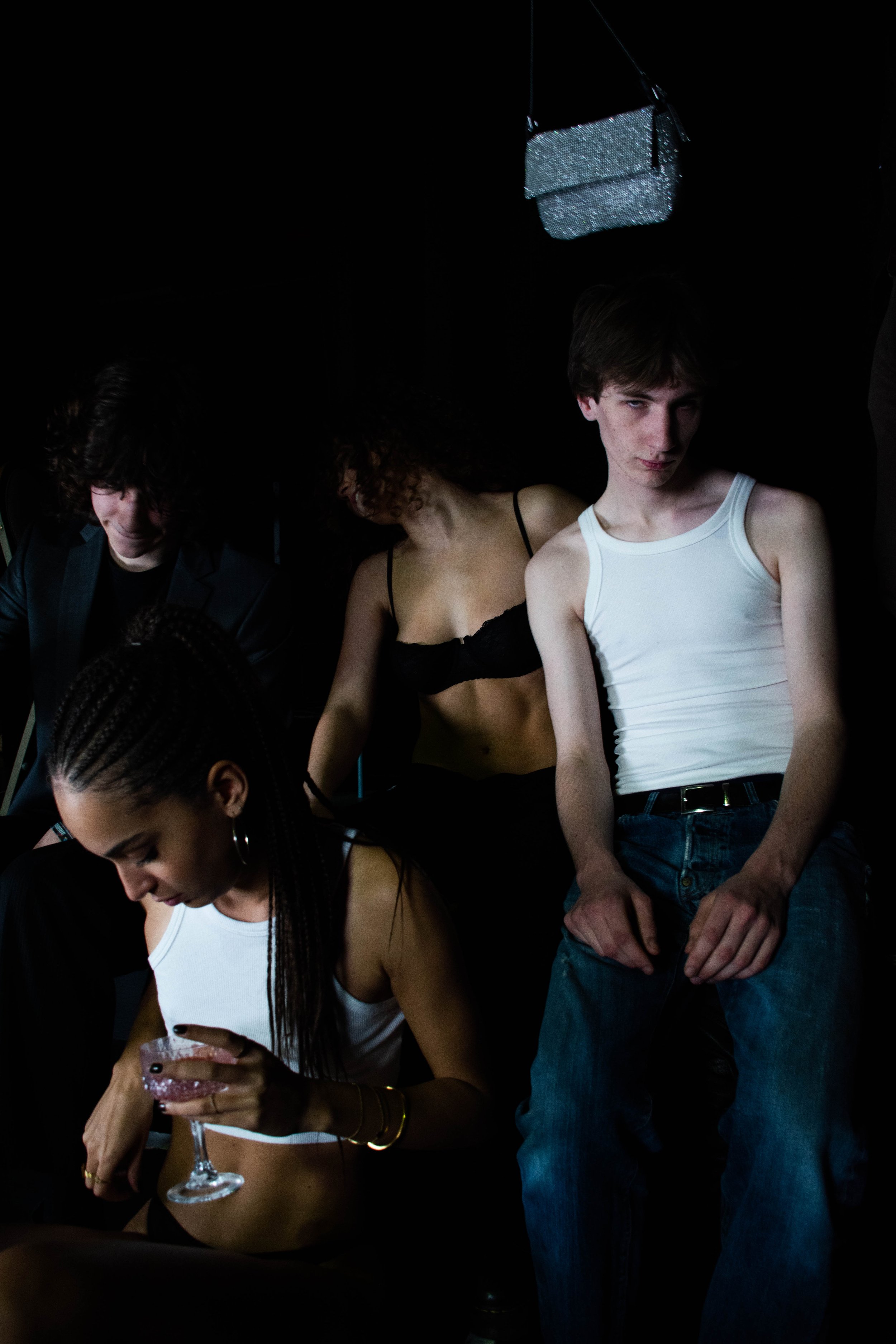 Group of young people in a dimly lit setting, with two women in white crop tops, one with braids holding a drink, and a young man in a white tank top leaning against the wall.