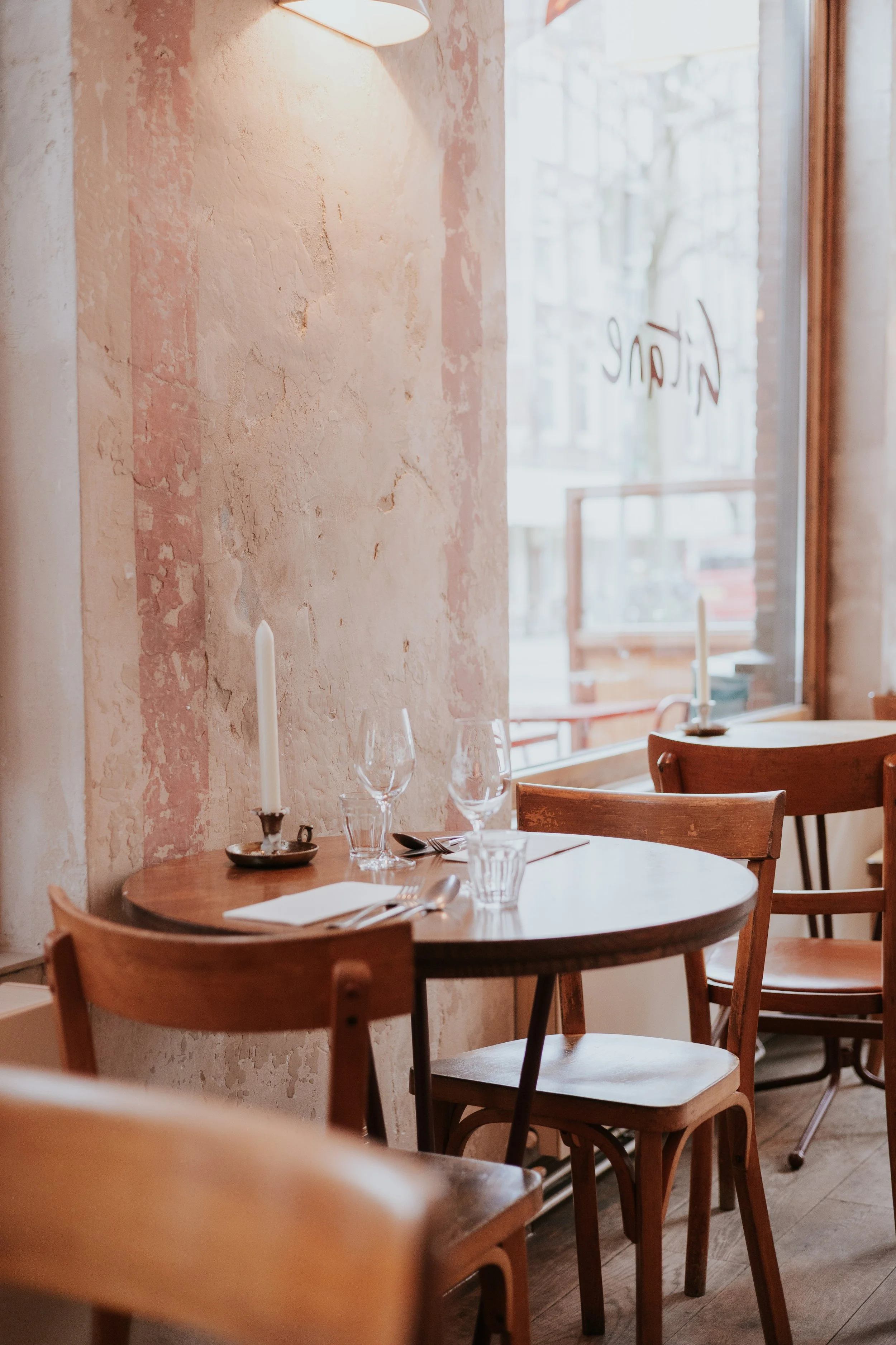A cozy restaurant corner with a round wooden table set with wine glasses, silverware, and a candle, situated near a large window with a sign that reads 'Nikon' in reverse.