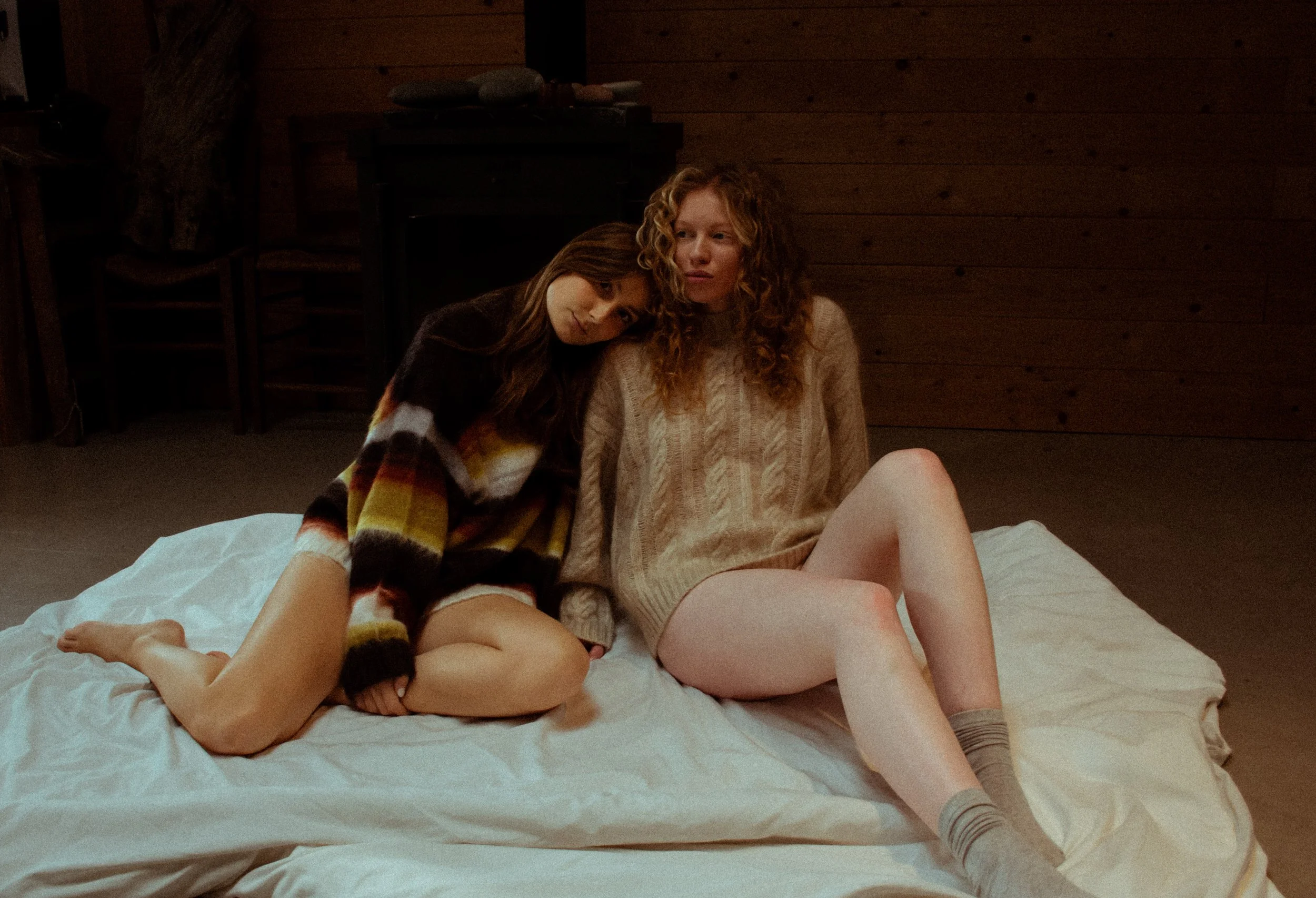 Two women sitting on a white bed in a room with wooden walls, one with curly red hair in a beige sweater and the other with long dark hair in a striped sweater, leaning on each other.