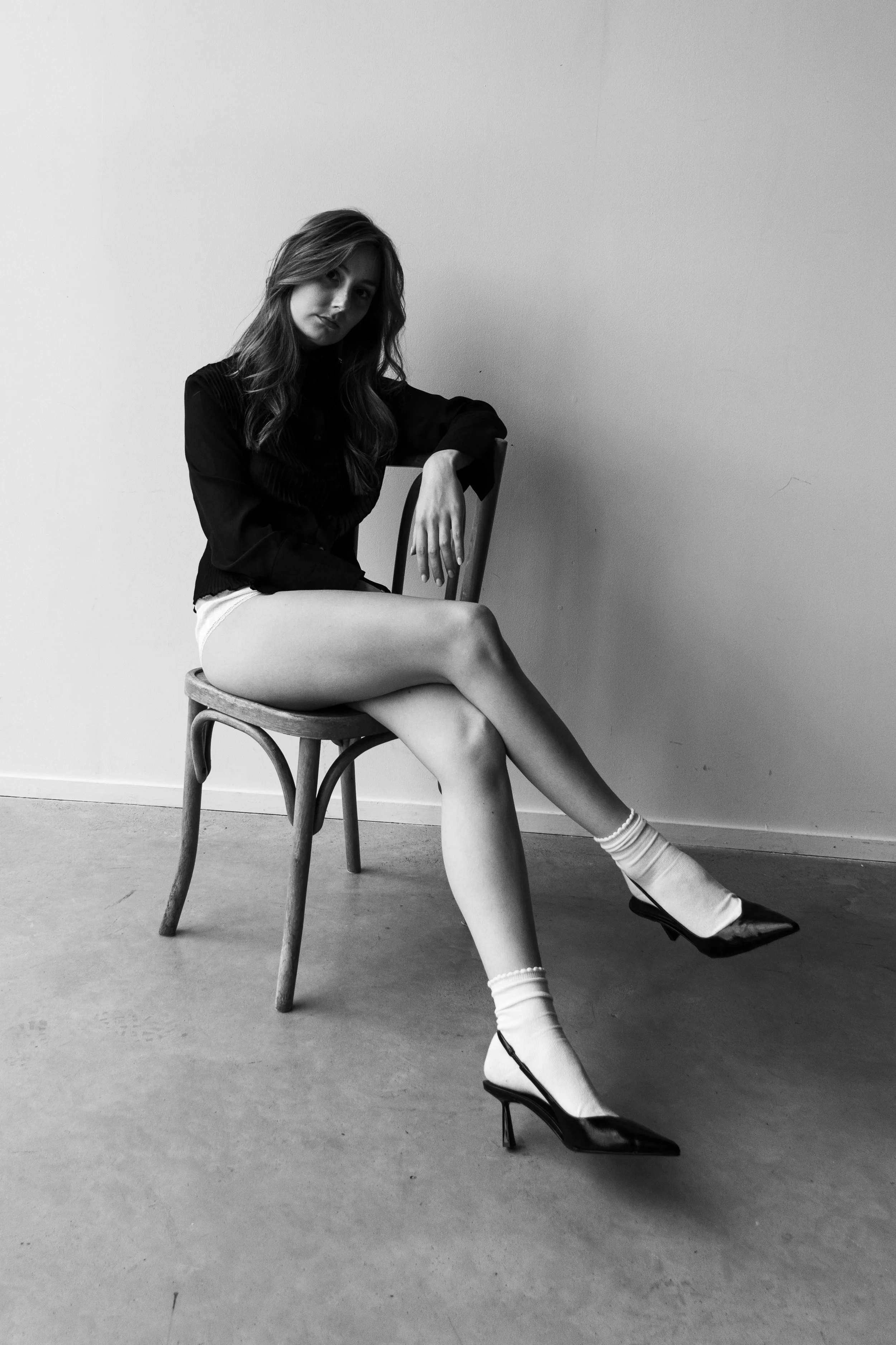 A woman with long hair sits on a wooden chair against a plain wall. She is wearing a dark top, light shorts, white socks, and black high heels. The image is in black and white.