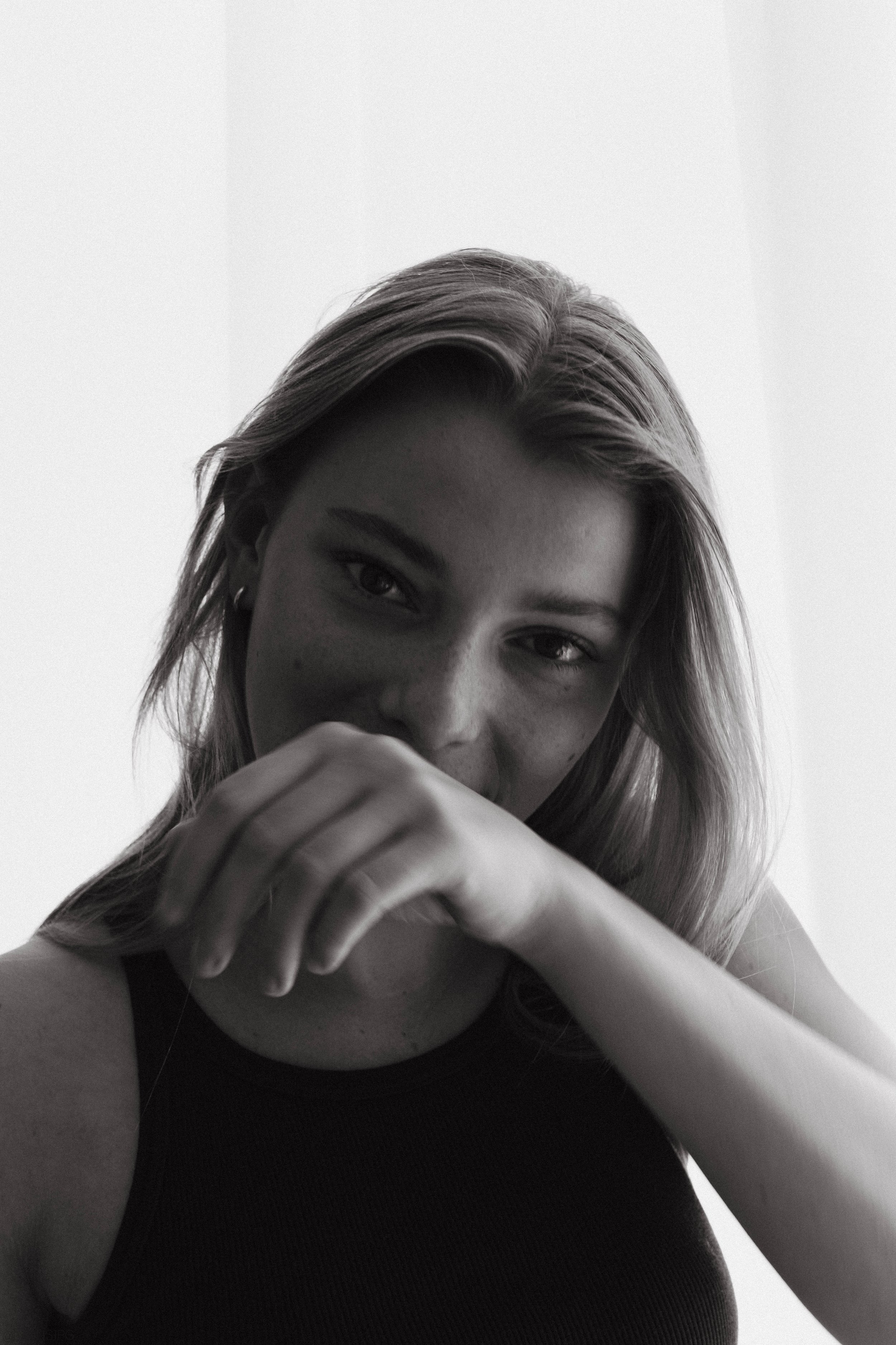 A black-and-white close-up portrait of a woman with light hair, looking at the camera, partially covering her face with her hand, against a plain white background.