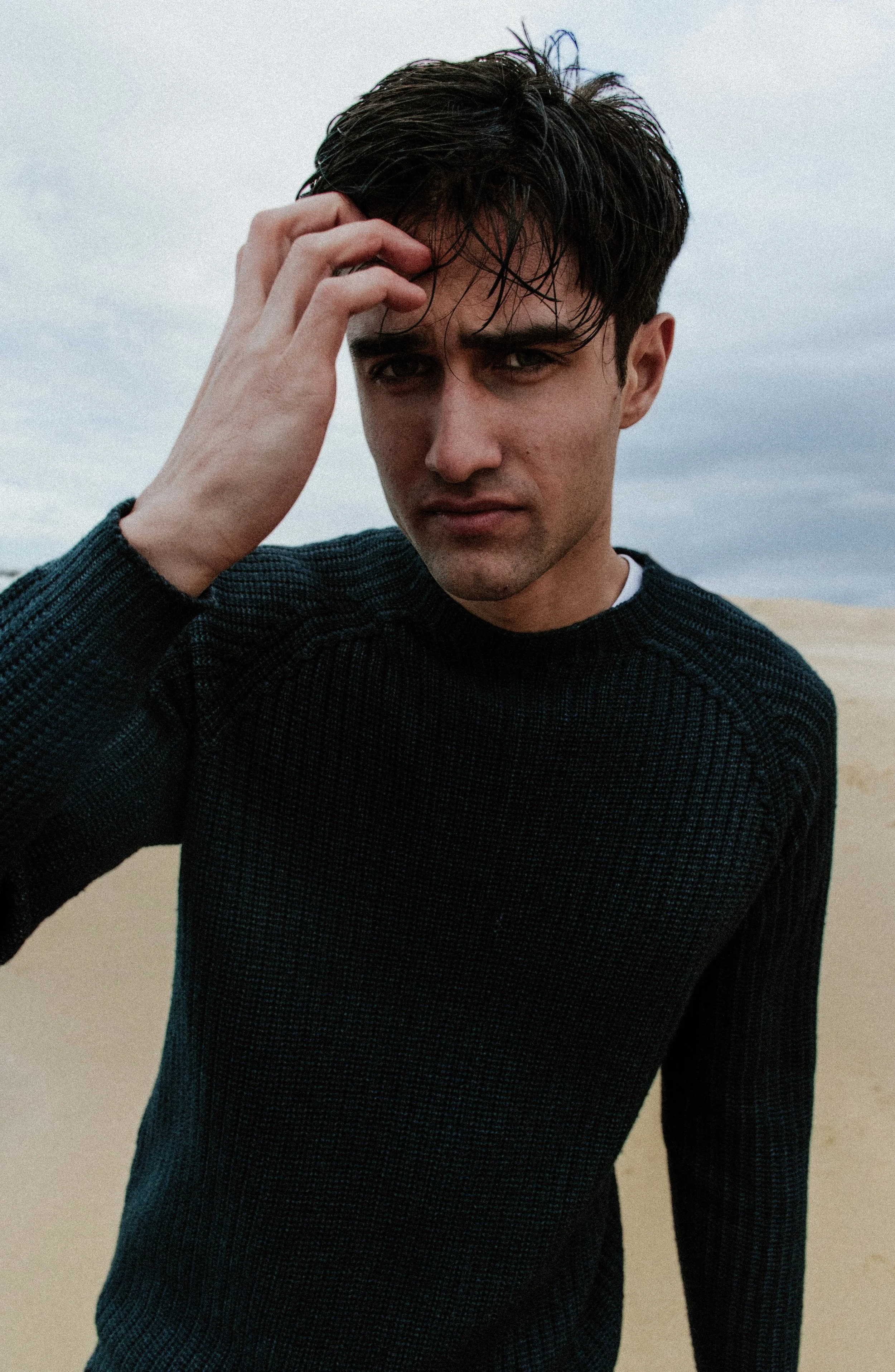 A young man with dark, wet hair in a black sweater, standing outdoors on a sandy landscape with a cloudy sky, looking into the camera with a serious expression.