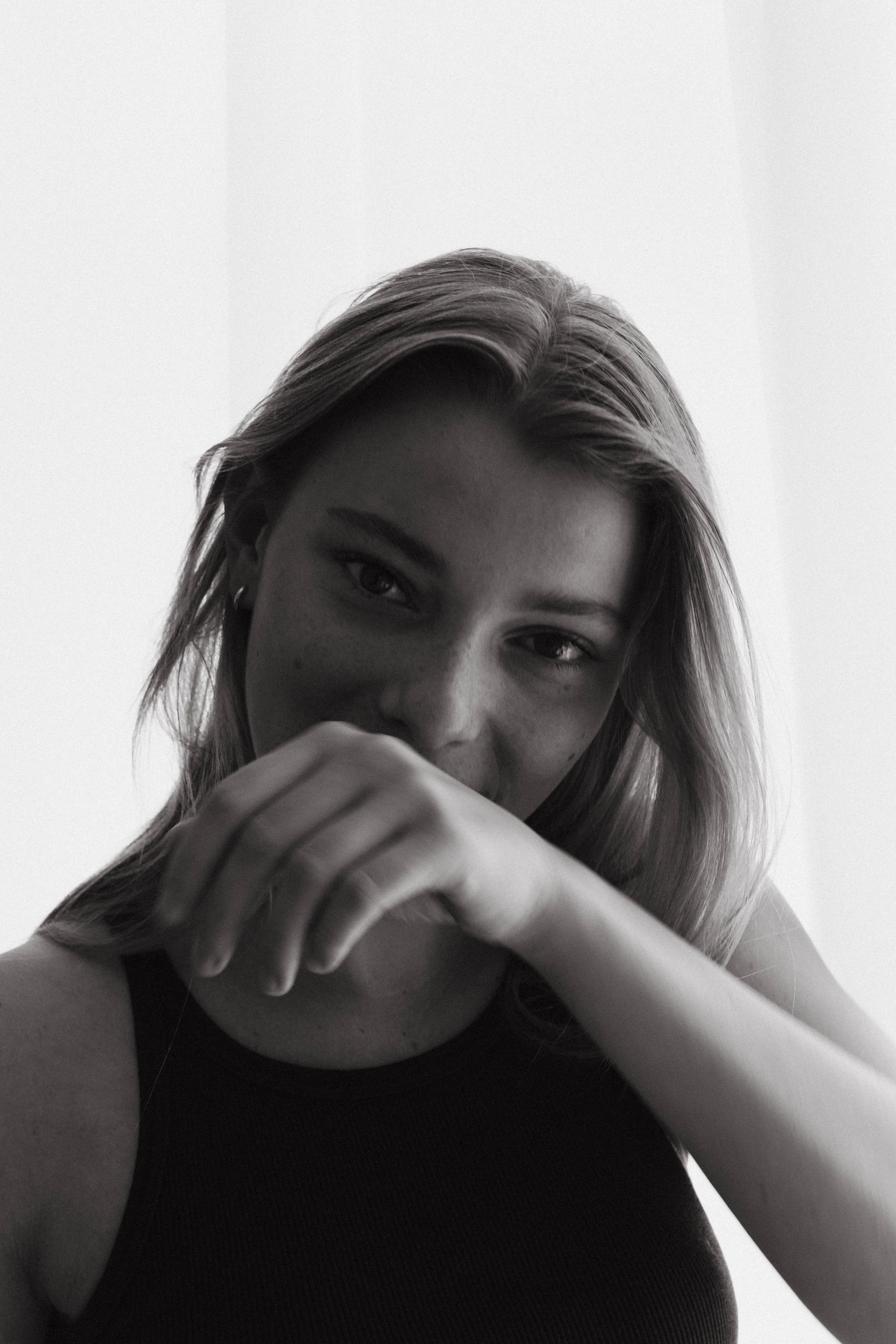 A black and white portrait of a woman with light hair, wearing a dark sleeveless top, resting her chin on her hand with a slight smile and direct gaze.