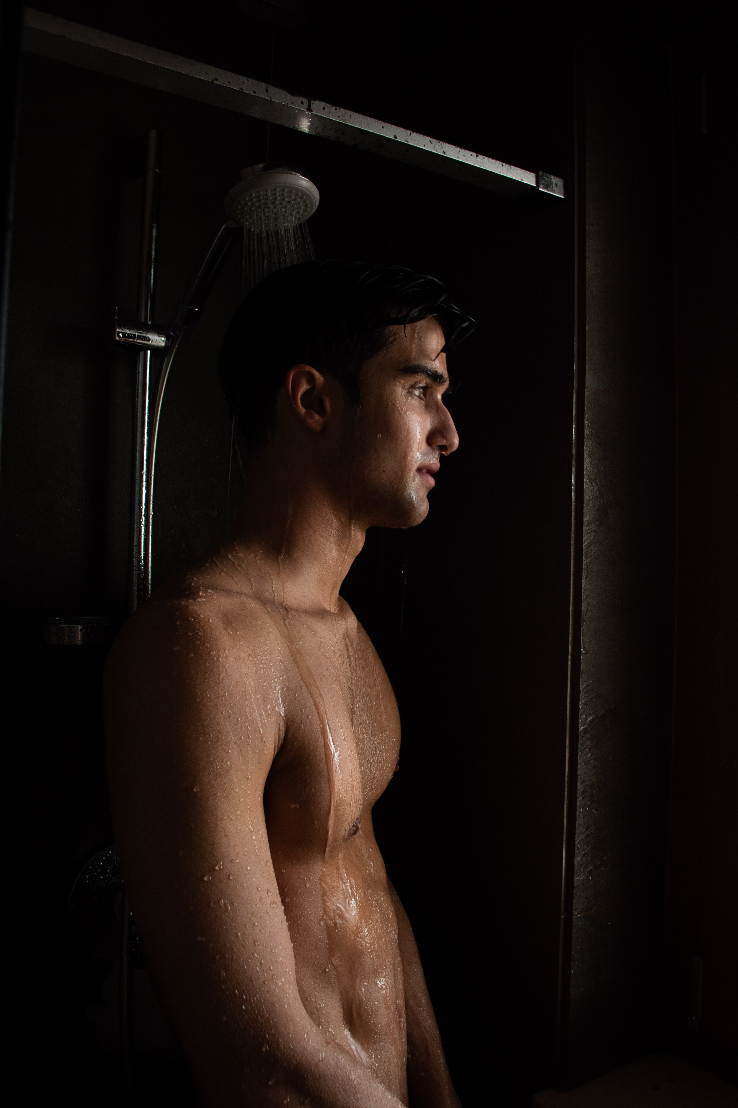 A shirtless man taking a shower with water running from a showerhead, standing against a dark background.