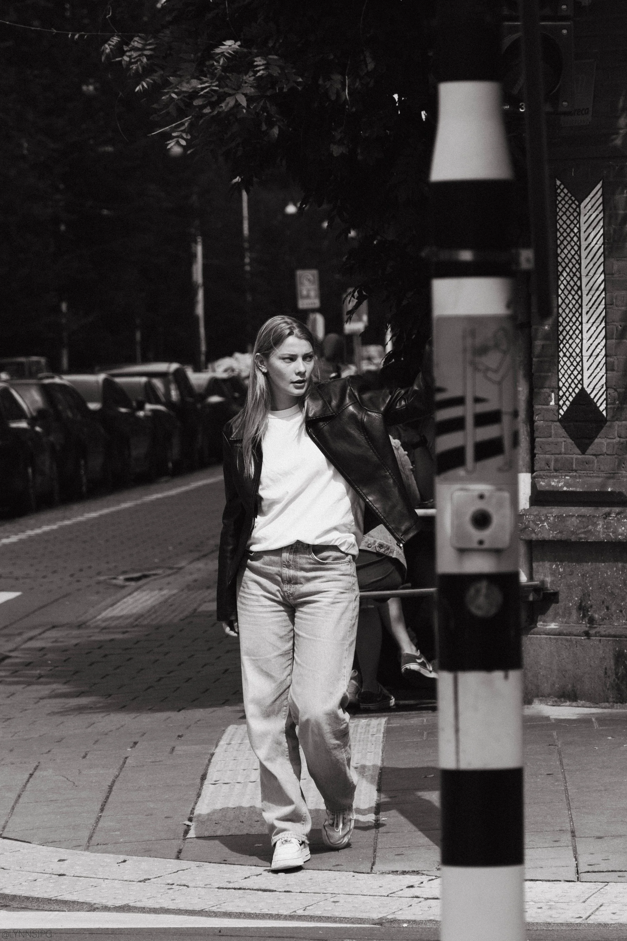 A young woman in a leather jacket, white t-shirt, and jeans is walking on a city sidewalk at night, holding onto a pole with her left hand.